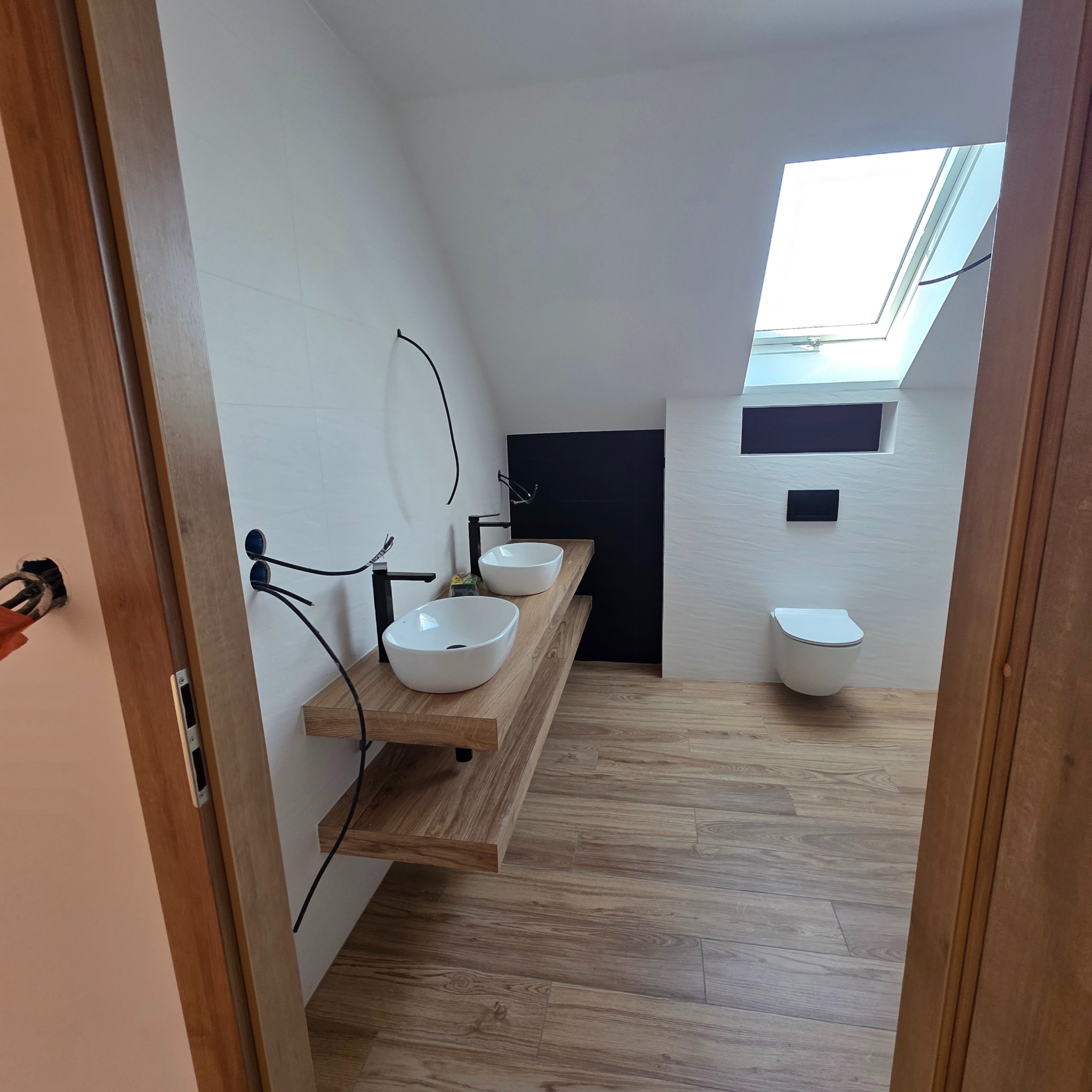 Nowoczesna łazienka na poddaszu z dwiema białymi umywalkami na drewnianym blacie, wiszącą toaletą i oknem dachowym. Widoczne niedokończone prace elektryczne.