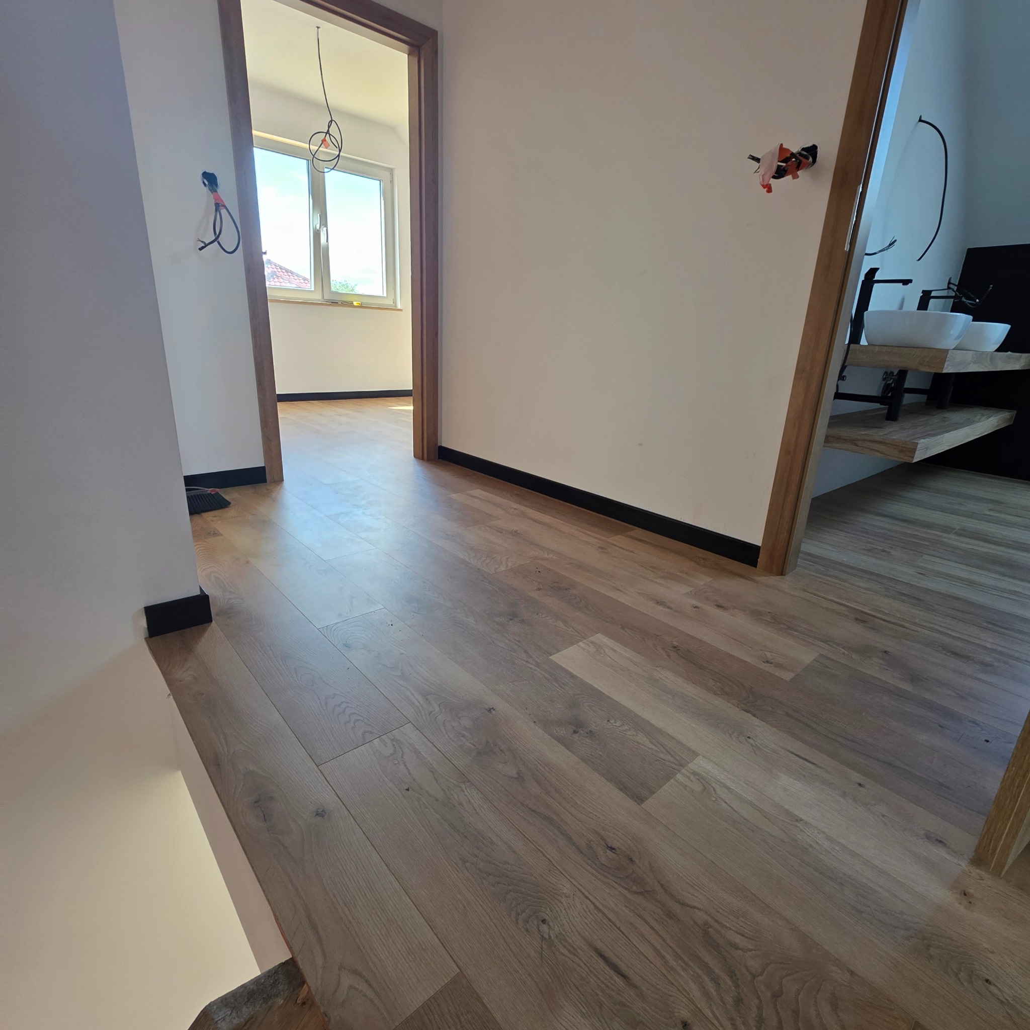 Jasne panele podłogowe w korytarzu z drewnianymi ościeżnicami i fragmentem łazienki z umywalkami na drewnianym blacie. Widoczne wystające kable elektryczne.