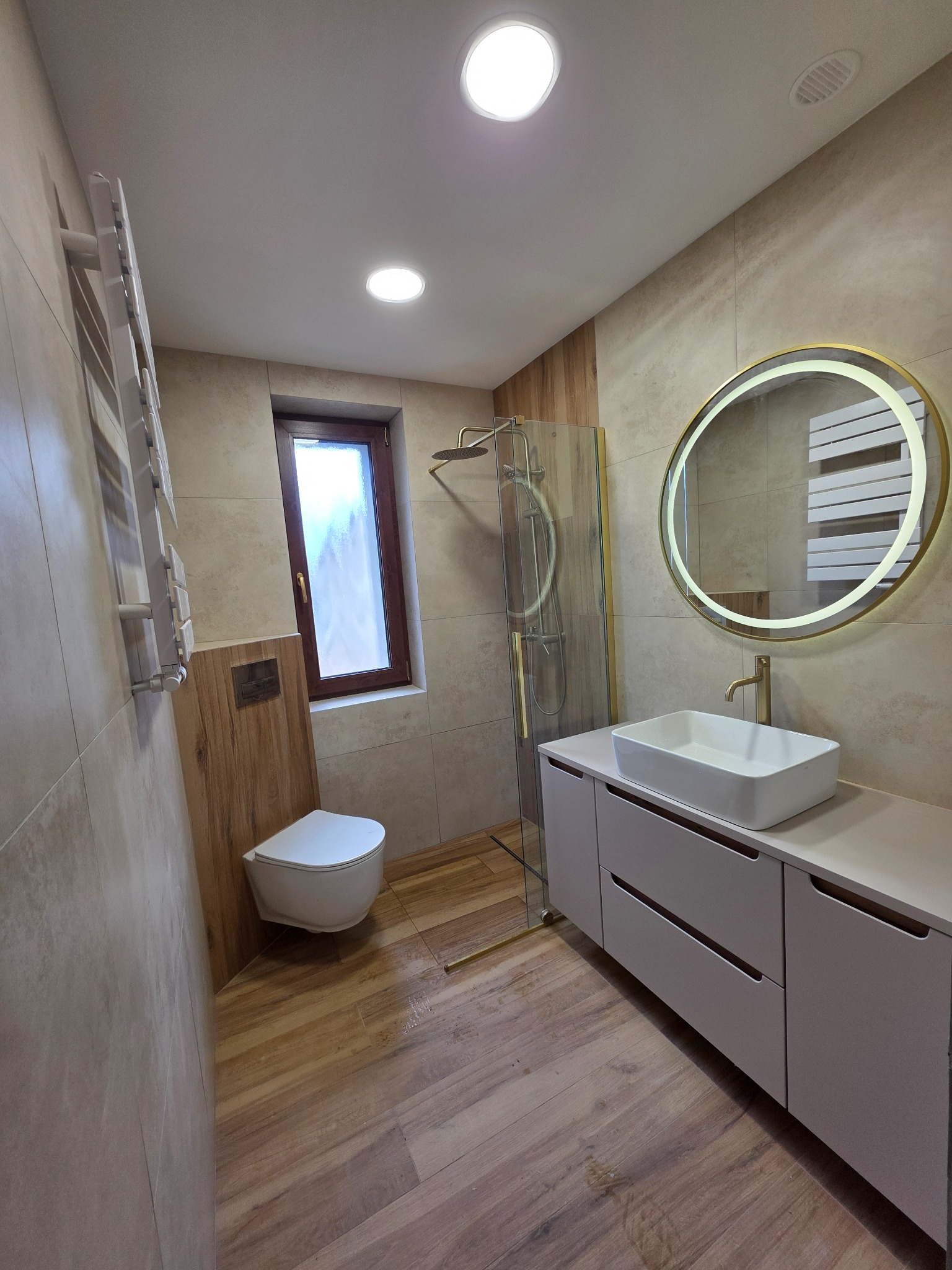 Nowoczesna łazienka z drewnopodobnymi płytkami, kabiną prysznicową ze złotymi detalami i okrągłym lustrem z podświetleniem LED. Umywalka nablatowa na szafce.