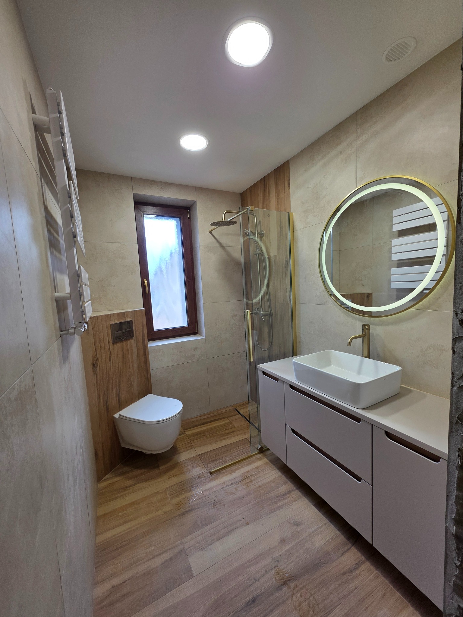 Nowoczesna łazienka z drewnopodobnymi płytkami, kabiną prysznicową ze złotymi detalami i okrągłym lustrem z podświetleniem LED. Umywalka nablatowa na szafce.