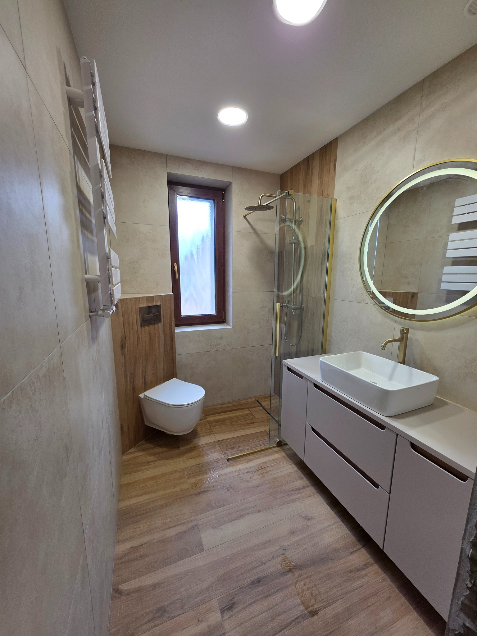 Nowoczesna łazienka z kabiną prysznicową, umywalką na szafce i okrągłym lustrem z podświetleniem LED. Uwagę przykuwają płytki imitujące drewno na podłodze i ścianie.