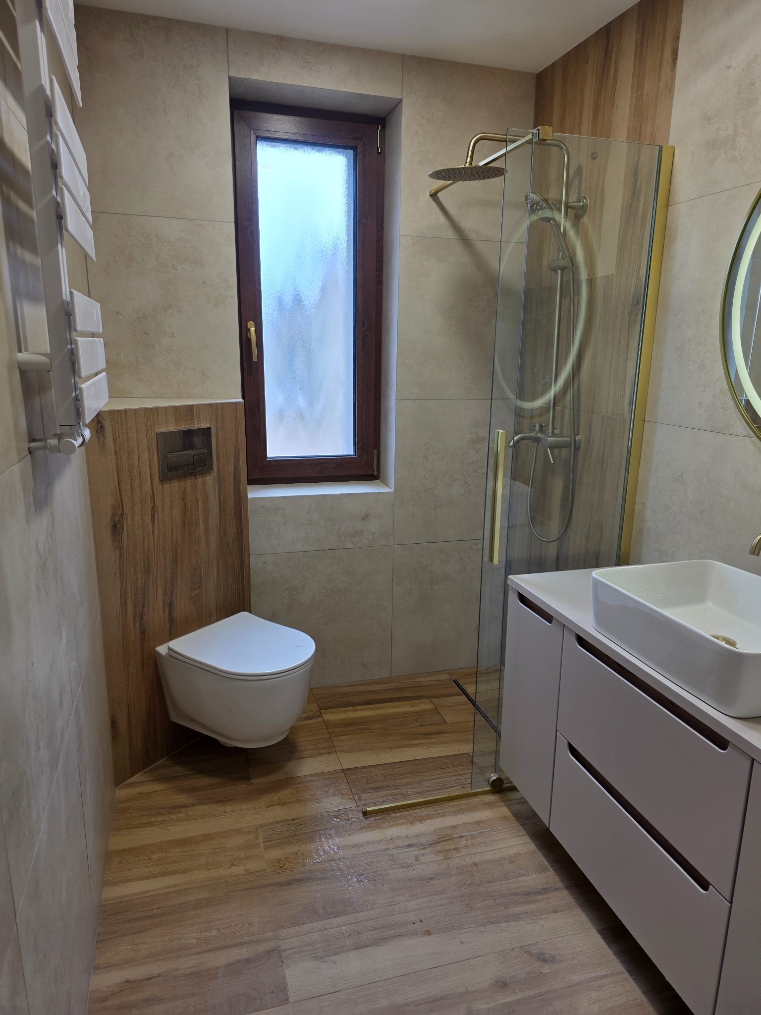 Nowoczesna łazienka po remoncie: beżowe płytki, drewnopodobna podłoga, kabina prysznicowa ze złotymi detalami, wisząca toaleta i szafka z umywalką.