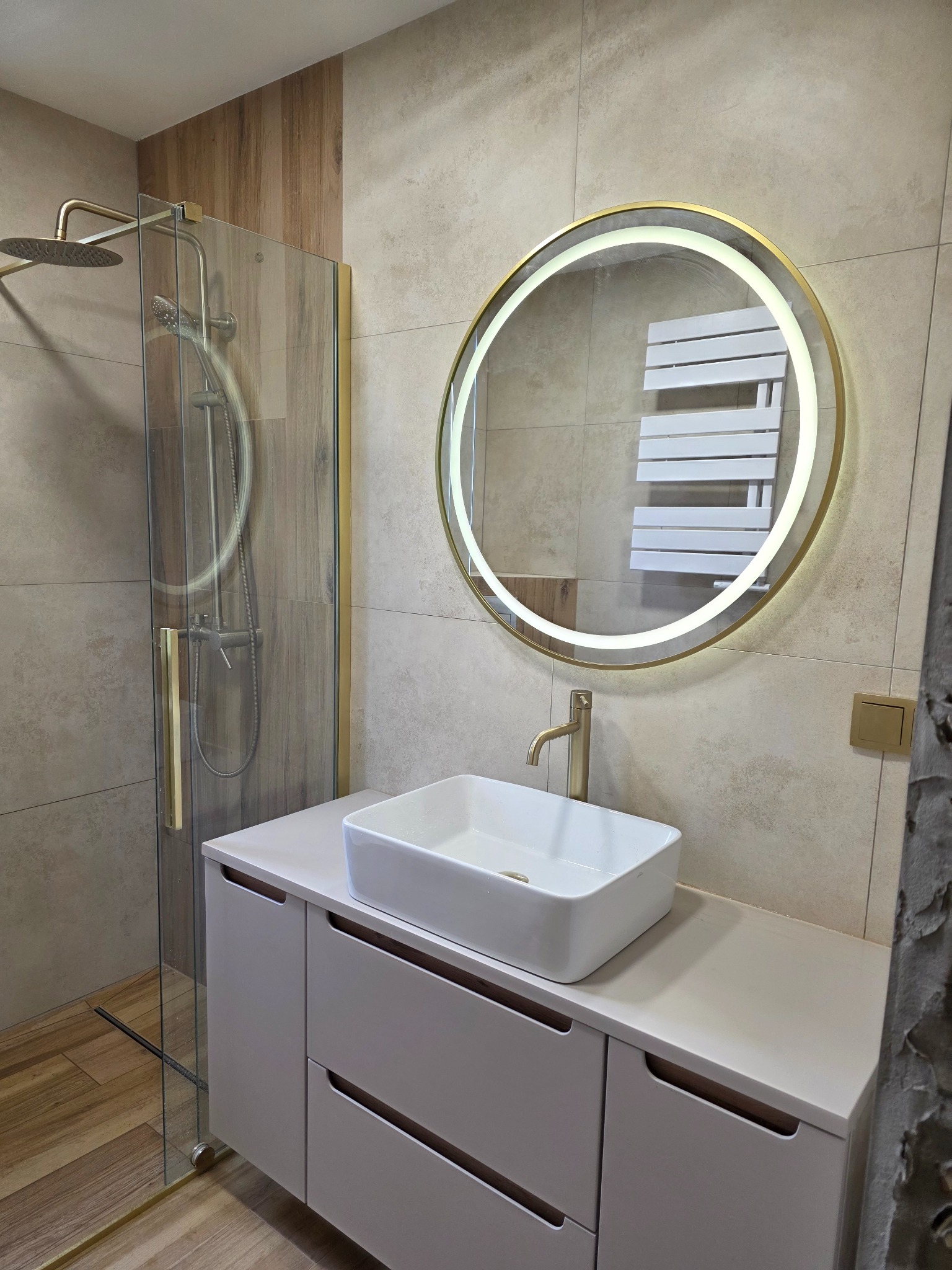 Nowoczesna łazienka z kabiną prysznicową typu walk-in, umywalką nablatową i okrągłym lustrem z podświetleniem LED. Beżowe płytki i drewniane akcenty.