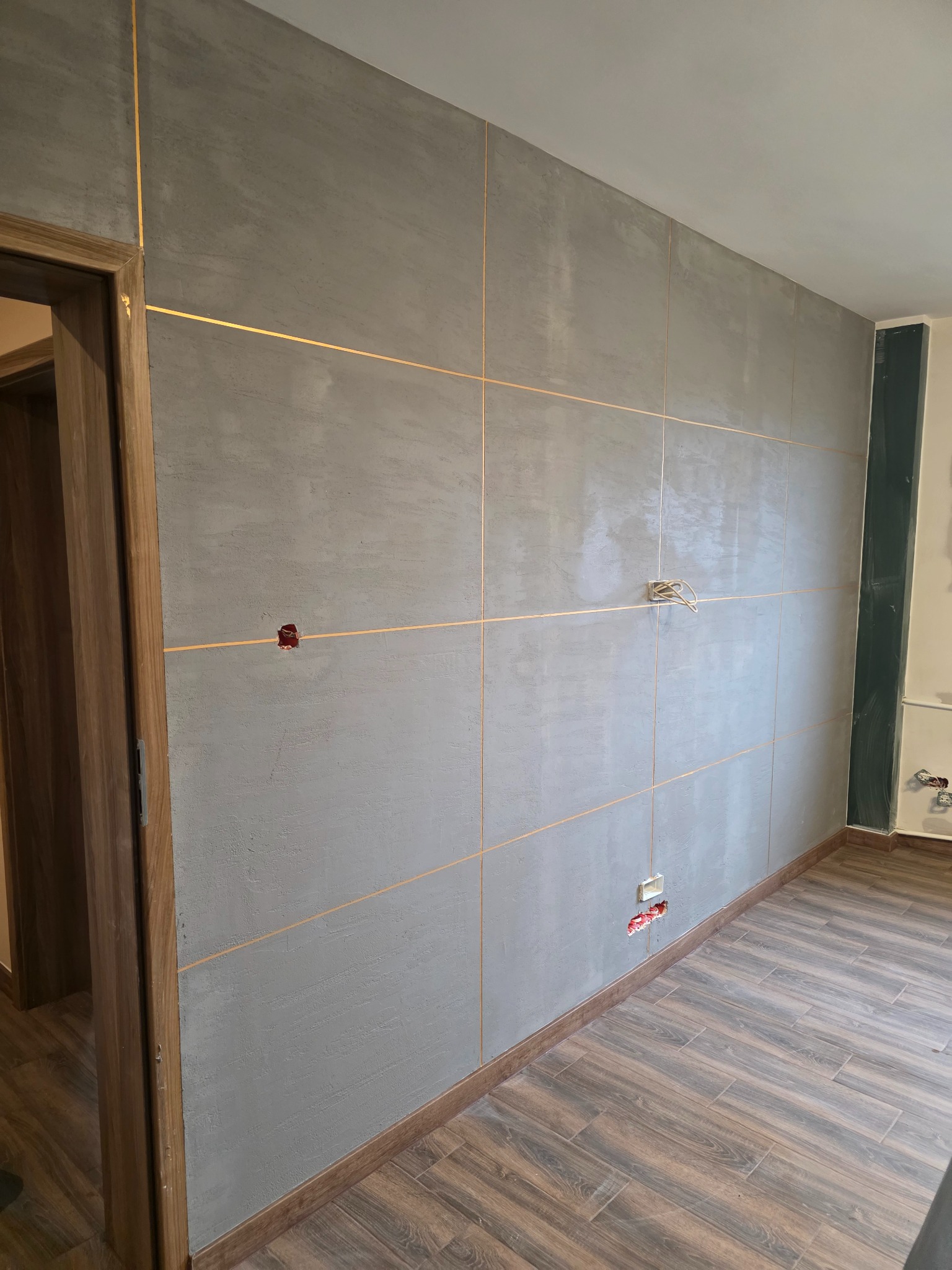 Ściana wykończona szarymi płytami z dekoracyjnymi, złotymi fugami. Widoczne gniazdka elektryczne i drewnopodobna podłoga. Fragment ościeżnicy drzwi po lewej stronie.