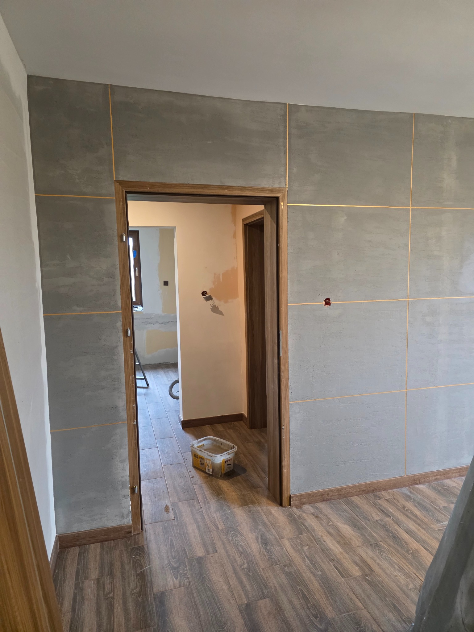 Wykończone wnętrze z szarymi płytami na ścianach, przedzielonymi złotymi listwami. Na podłodze panele imitujące drewno. Widoczne drzwi i fragment pomieszczenia w głębi.