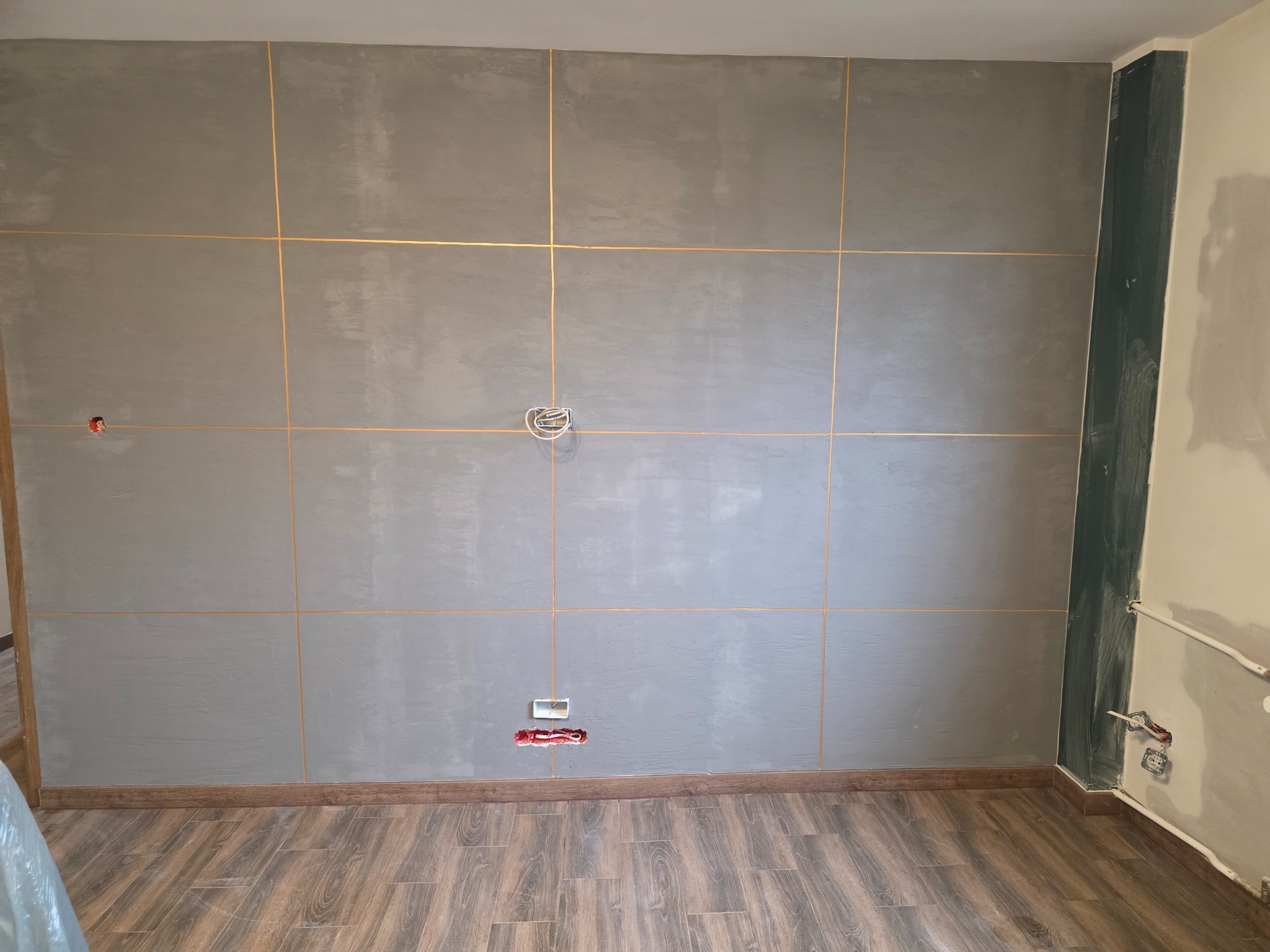Ściana w szarym odcieniu z dekoracyjnymi, złotymi liniami imitującymi płytki. Widoczne gniazdka elektryczne i drewnopodobna podłoga. Wykończenie wnętrza w trakcie prac.