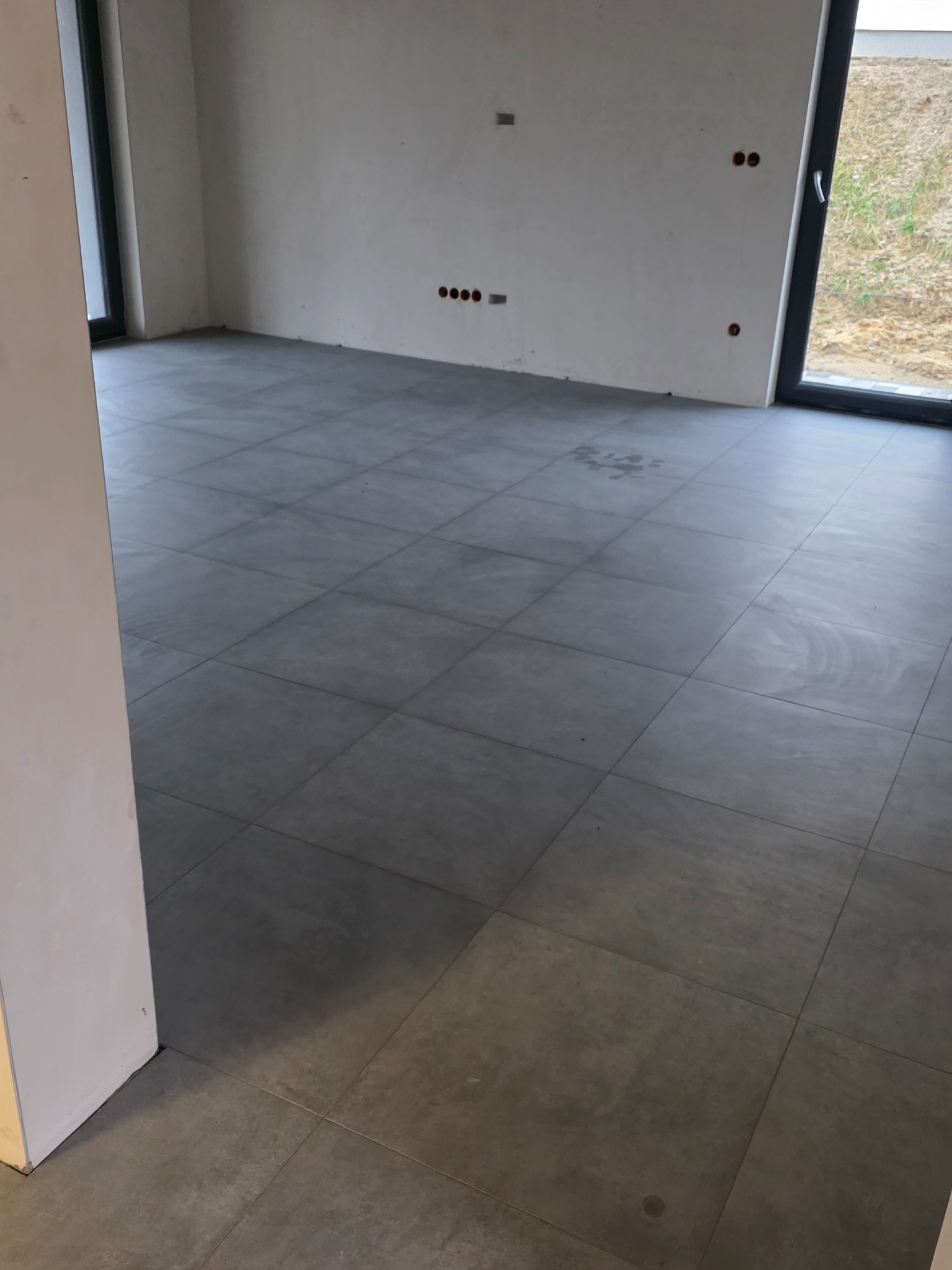 Szare, kwadratowe płytki podłogowe ułożone w pustym pomieszczeniu z białymi ścianami i widocznymi puszkami elektrycznymi. Widok z perspektywy podłogi.