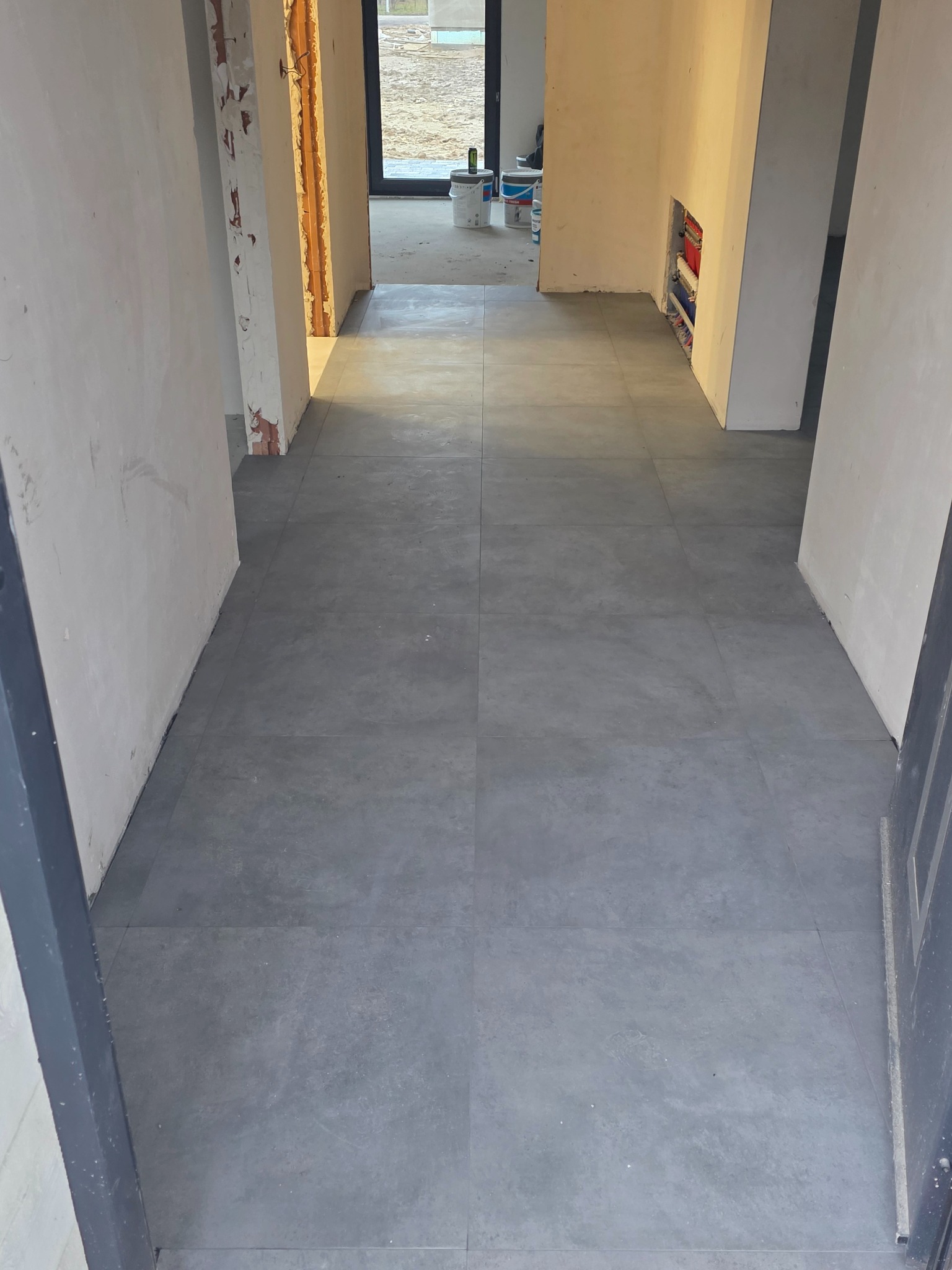 Ułożone szare płytki podłogowe w korytarzu w trakcie remontu. Widoczne ściany w stanie surowym i instalacje. W tle okno i puszki z farbą.