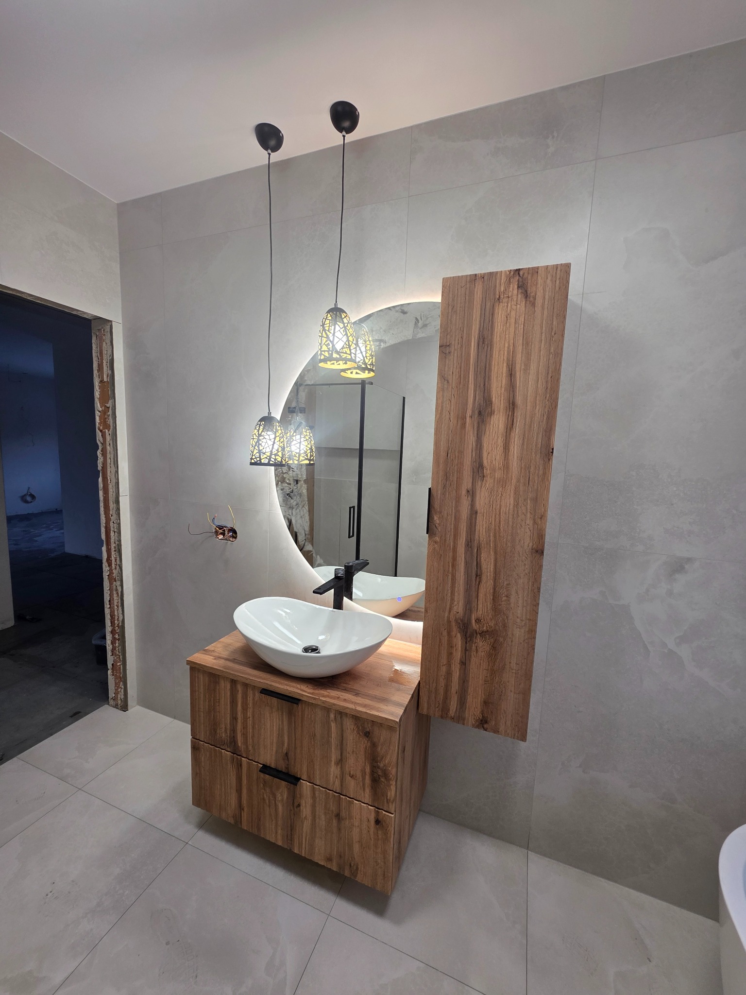 Nowoczesna łazienka z drewnianą szafką pod umywalką, okrągłym lustrem z podświetleniem LED i szarymi płytkami imitującymi marmur. Widoczne wiszące lampy i fragment kabiny prysznicowej.