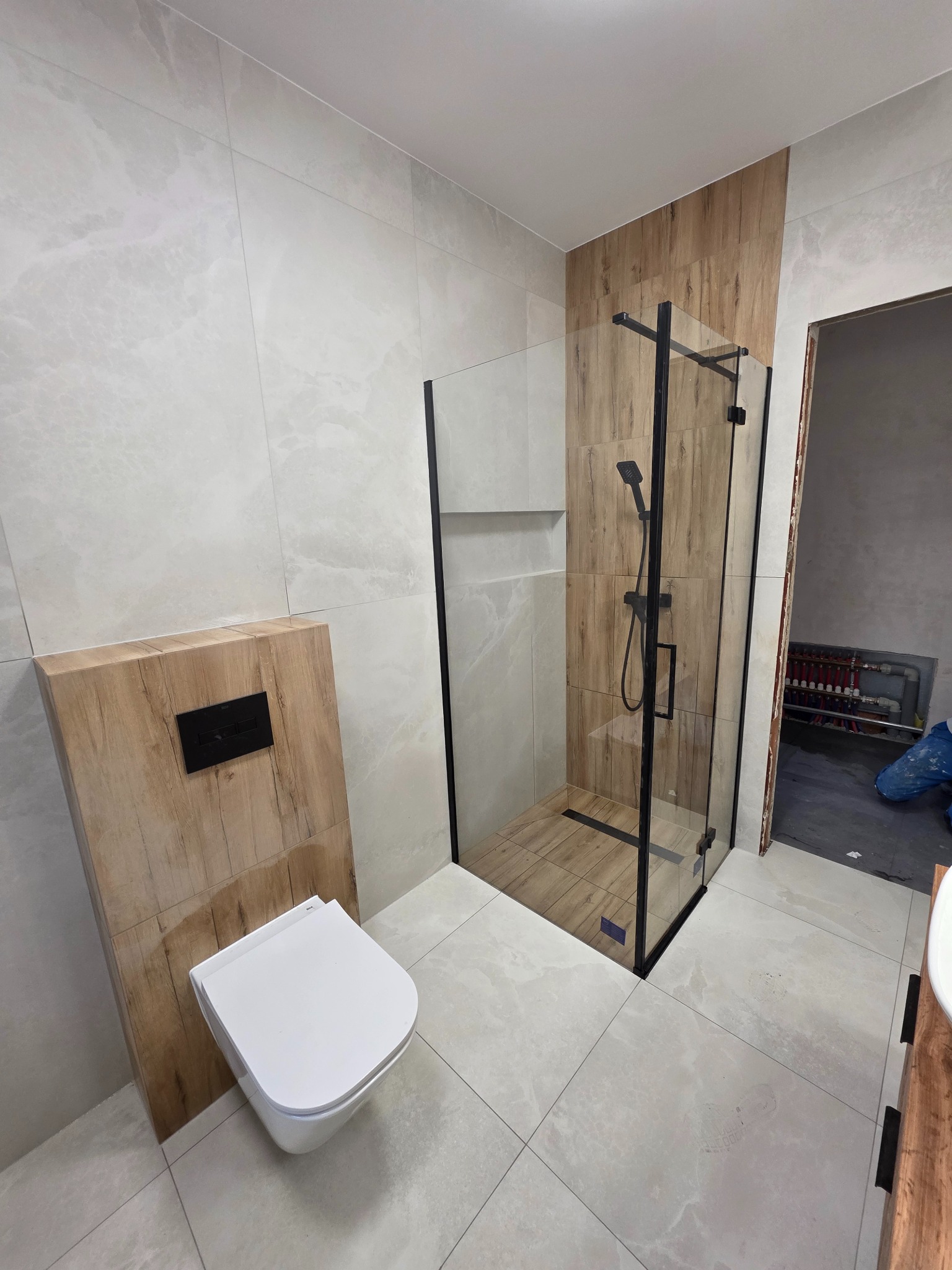 Nowoczesna łazienka z kabiną prysznicową typu walk-in z czarnymi ramami, podwieszaną toaletą i płytkami imitującymi drewno. Widoczne płytki w odcieniach szarości.