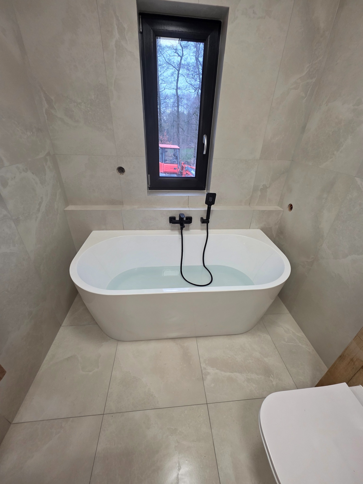 Nowoczesna łazienka z wolnostojącą wanną wypełnioną wodą, wykończona płytkami w odcieniach beżu. W tle okno z widokiem na las i czerwony traktor.