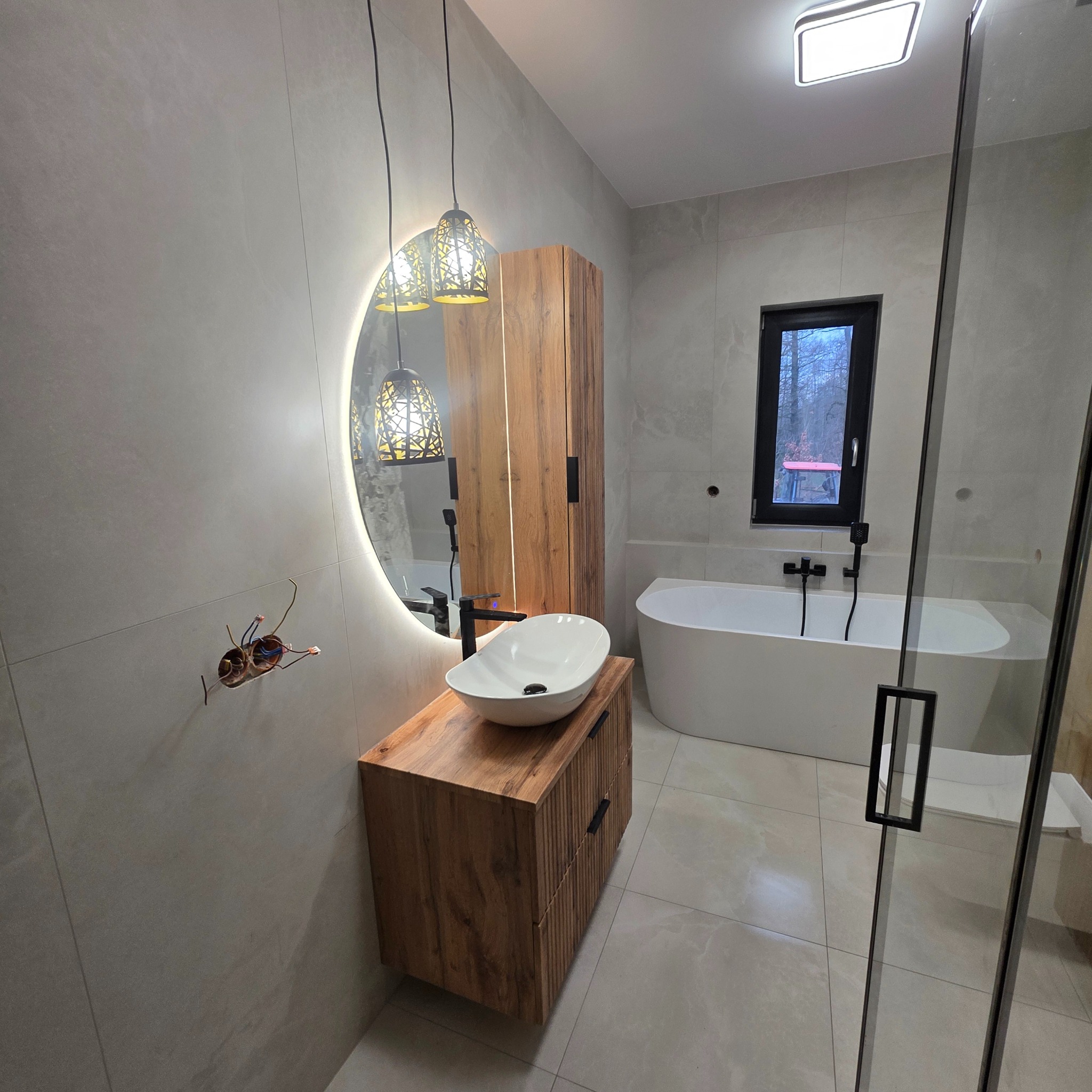 Nowoczesna łazienka z drewnianą szafką pod umywalkę, okrągłym lustrem z podświetleniem LED i wolnostojącą wanną przy oknie. Szare płytki na ścianach i podłodze.