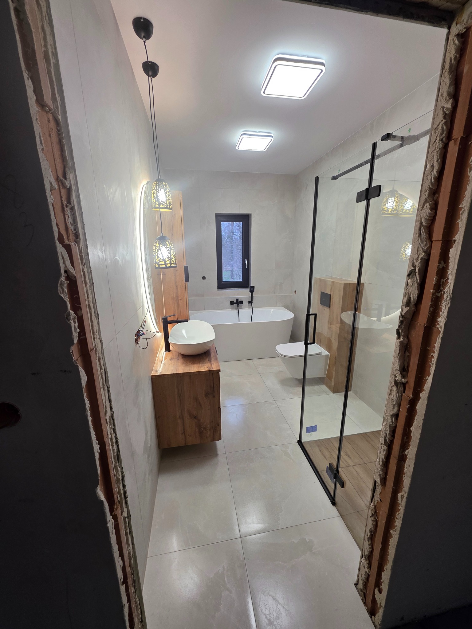 Nowoczesna łazienka z kabiną prysznicową typu walk-in, umywalką na drewnianej szafce i podświetlanym lustrem, widziana zza surowego otworu drzwiowego.