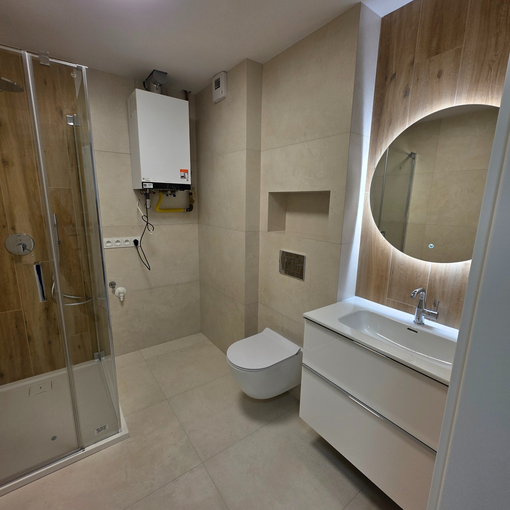 Jasna łazienka po remoncie: kabina prysznicowa, podwieszana toaleta, szafka z umywalką i okrągłe lustro z podświetleniem. Na ścianie piec gazowy.