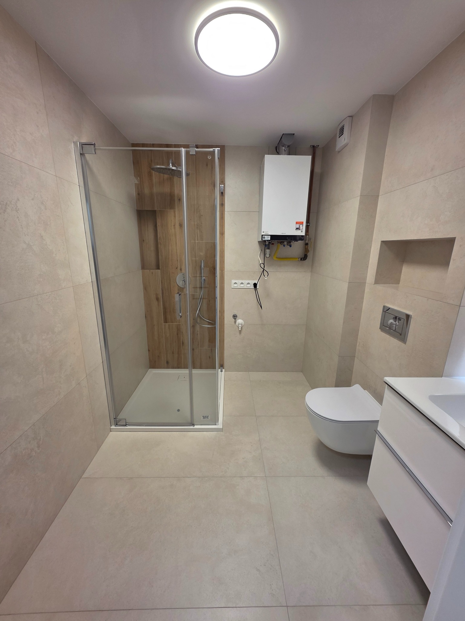 Jasna łazienka po remoncie: kabina prysznicowa ze szklanymi drzwiami, wisząca toaleta, umywalka z szafką. Na ścianie piec gazowy i wnęka. Płytki w odcieniach beżu.