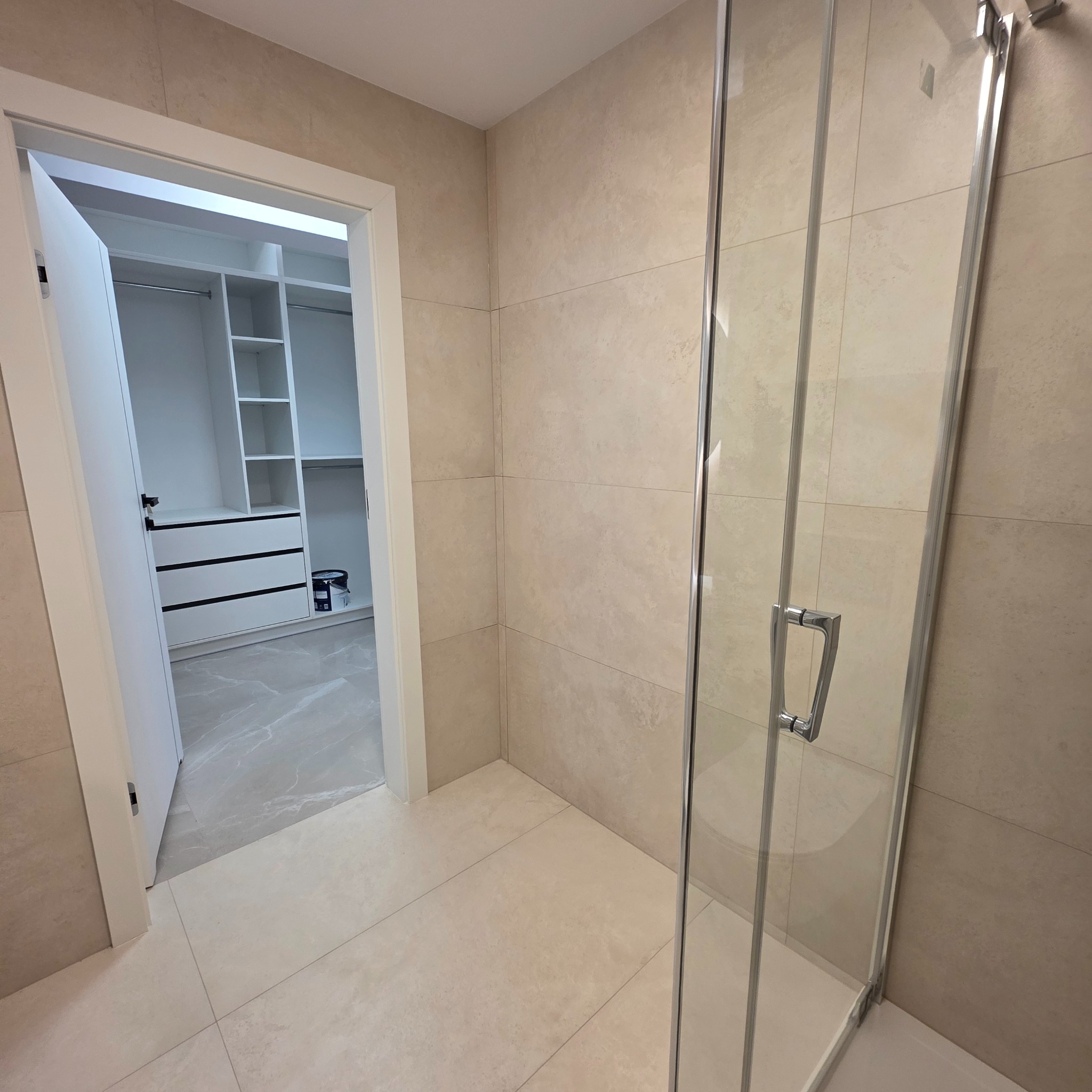 Beżowa łazienka z kabiną prysznicową i wejściem do garderoby z białymi meblami. Widoczne płytki na ścianach i podłodze, szklane drzwi kabiny.