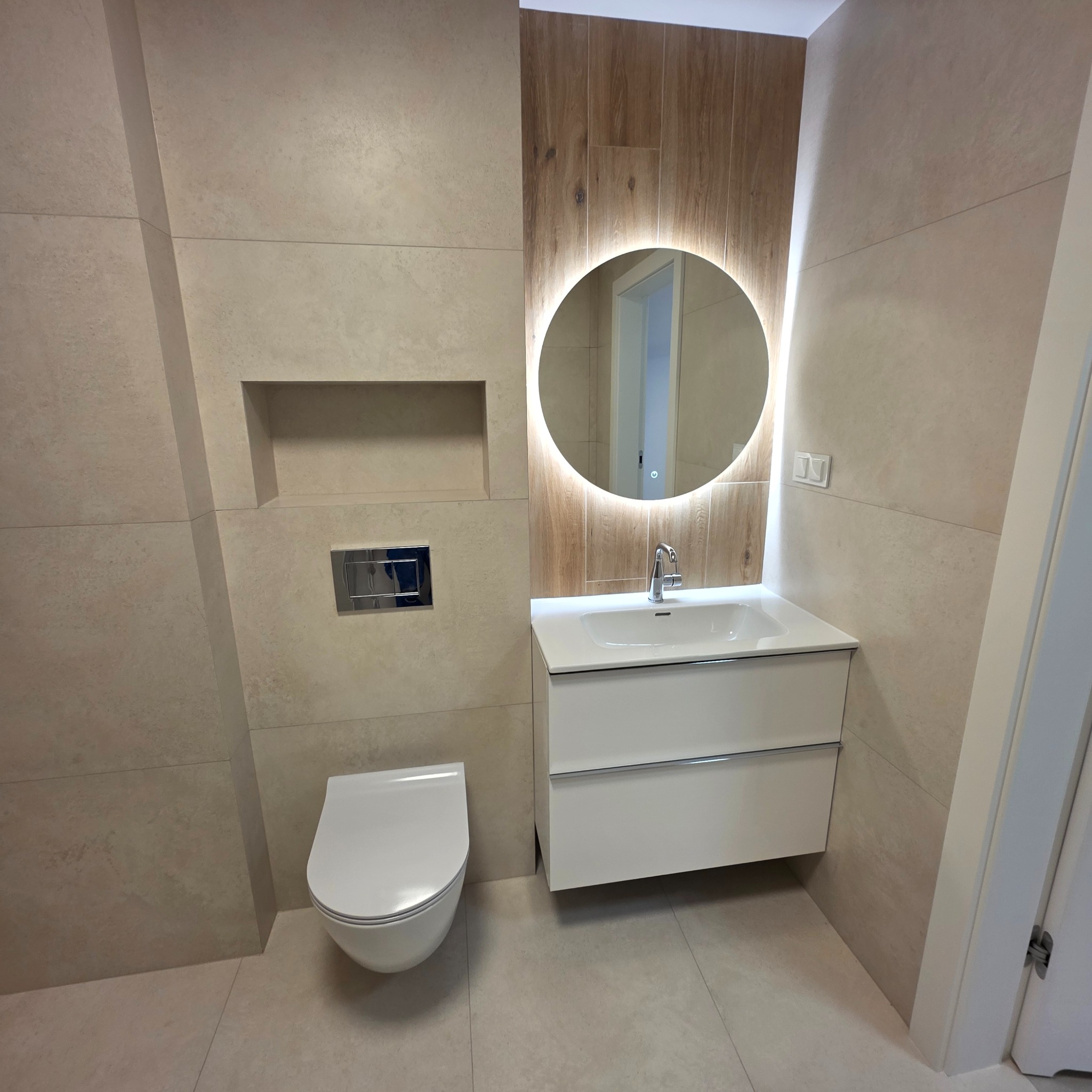 Nowoczesna łazienka z beżowymi płytkami, podświetlanym lustrem i wiszącą toaletą. Minimalistyczny design z drewnianym akcentem za lustrem.