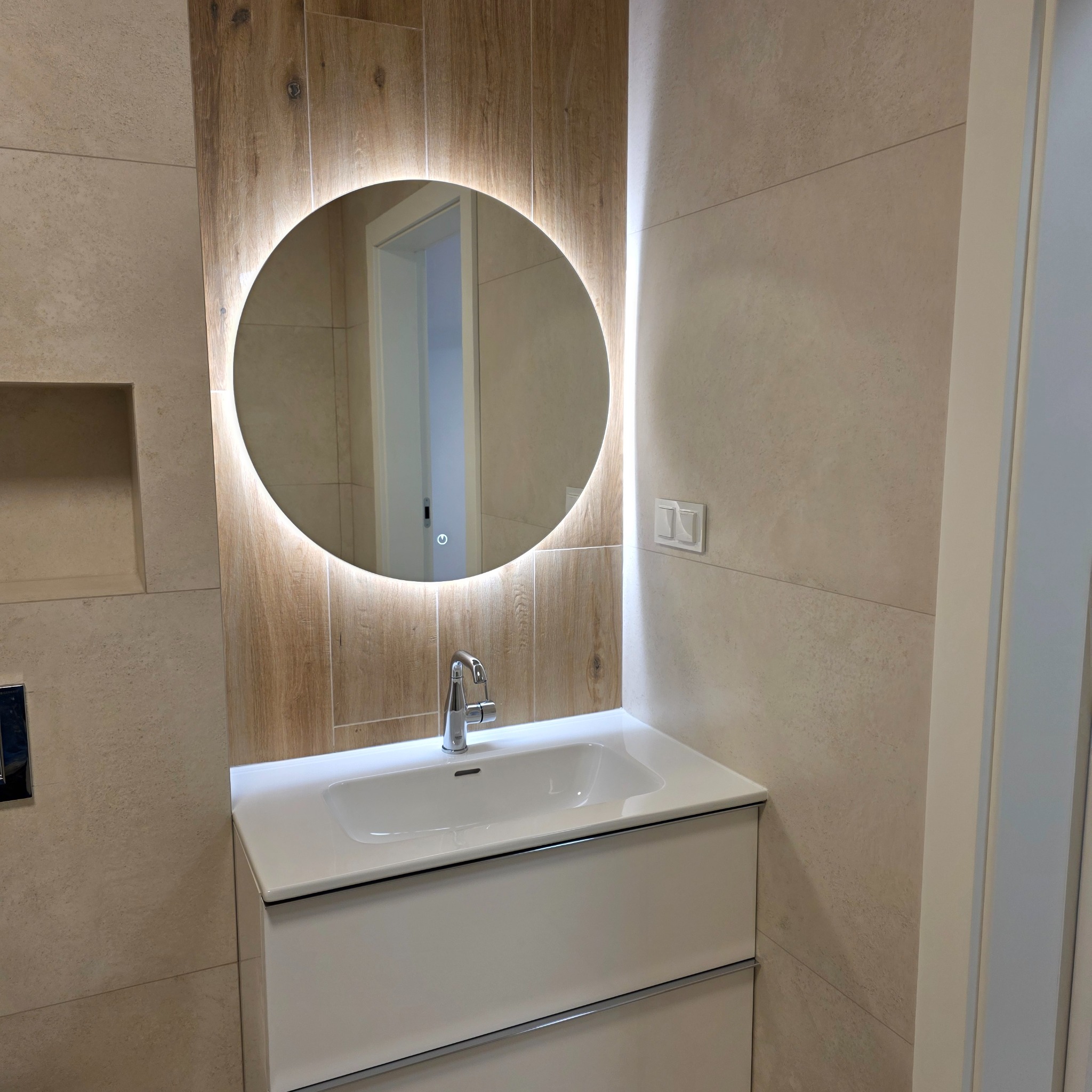 Nowoczesna łazienka po remoncie: beżowe płytki, okrągłe lustro z podświetleniem LED, biała szafka z umywalką i chromowana bateria. Widoczna wnęka w ścianie.