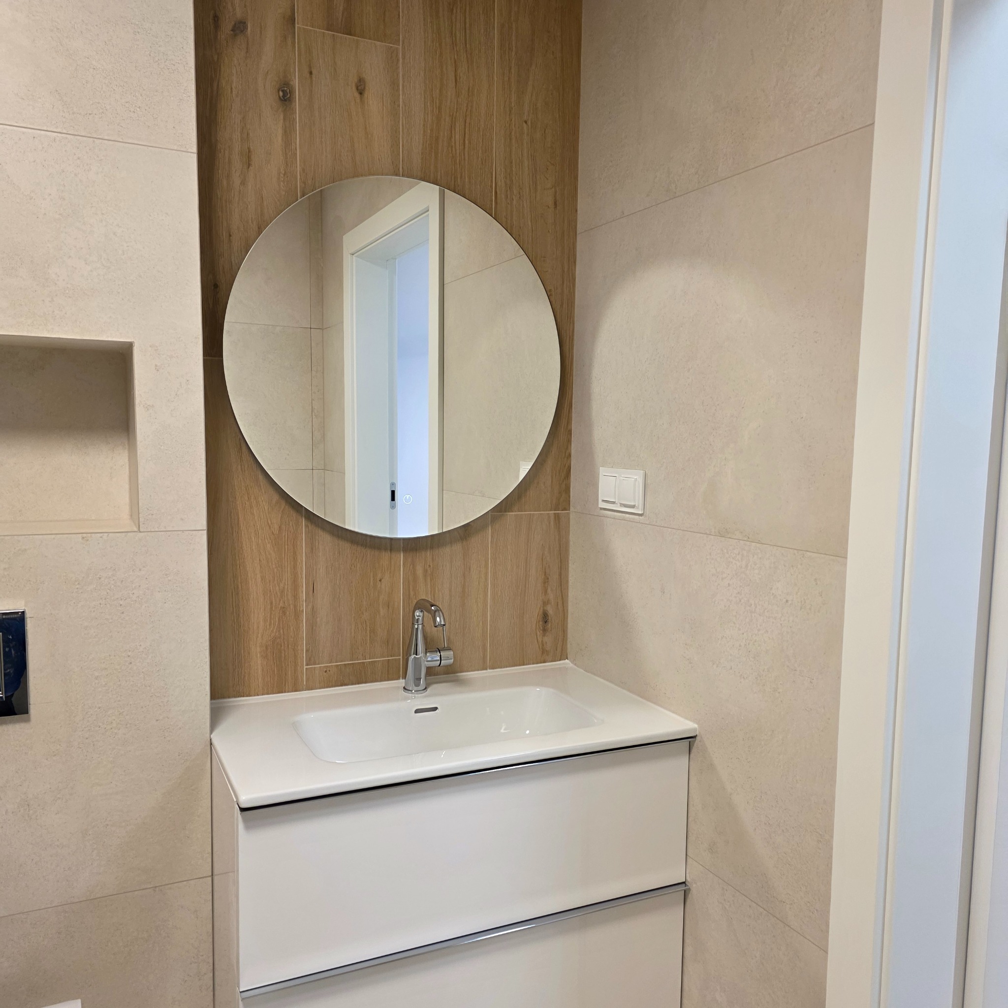 Nowoczesna łazienka z owalnym lustrem, białą szafką pod umywalką i beżowymi płytkami imitującymi kamień oraz drewno. Minimalistyczny design, chromowana bateria.