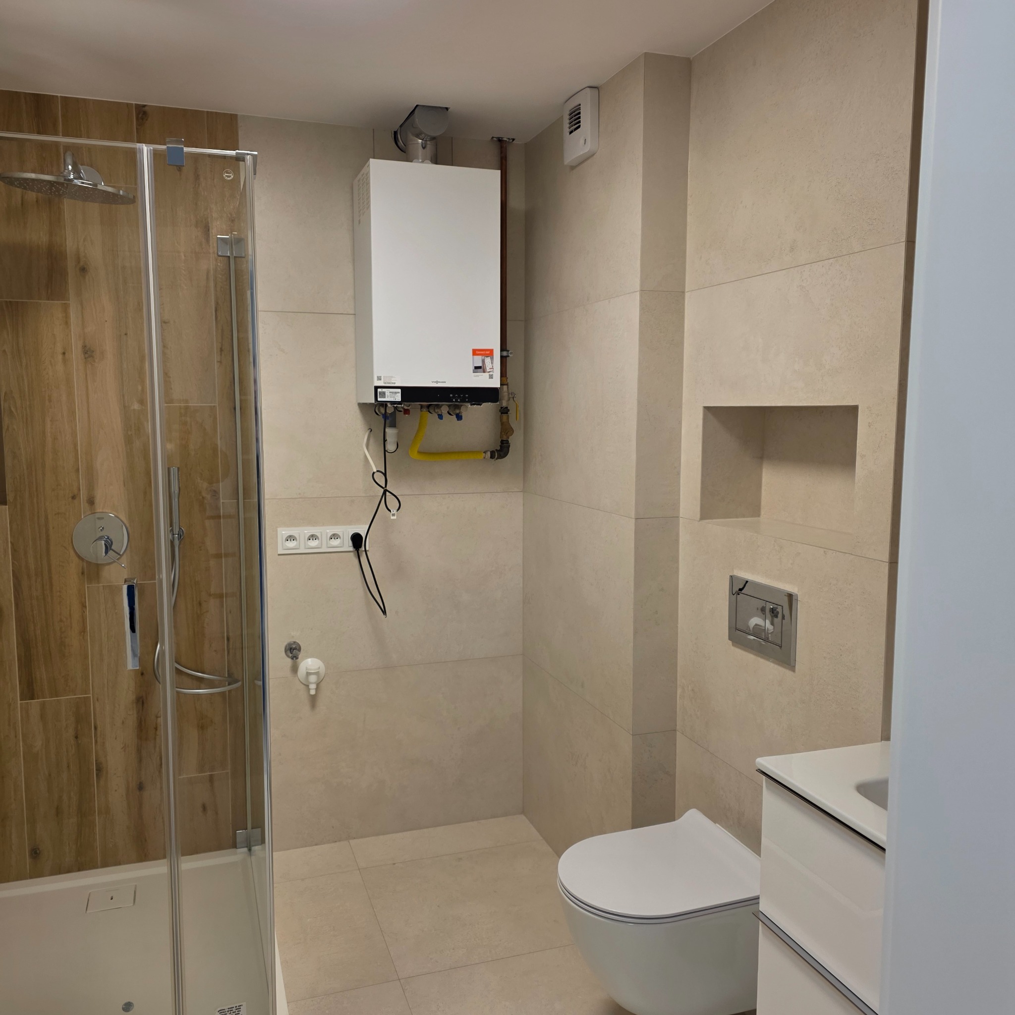 Nowoczesna łazienka po remoncie: kabina prysznicowa z drewnopodobnymi płytkami, beżowe płytki na ścianach, podwieszana toaleta i piec gazowy na ścianie.