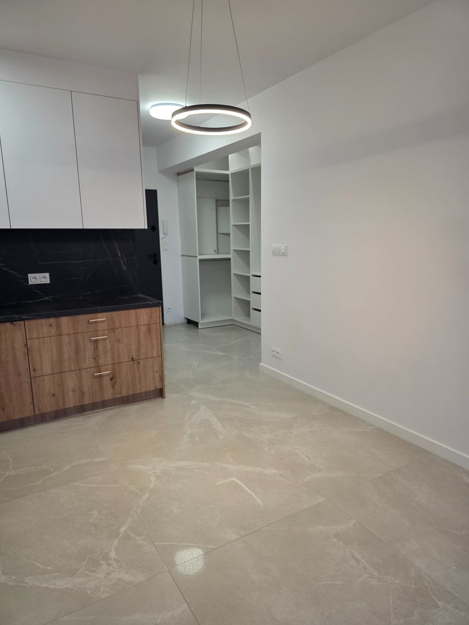 Nowoczesne wnętrze: beżowe płytki podłogowe, drewniana zabudowa kuchenna z czarnym blatem i białe szafy wnękowe. Okrągła lampa sufitowa dodaje elegancji.