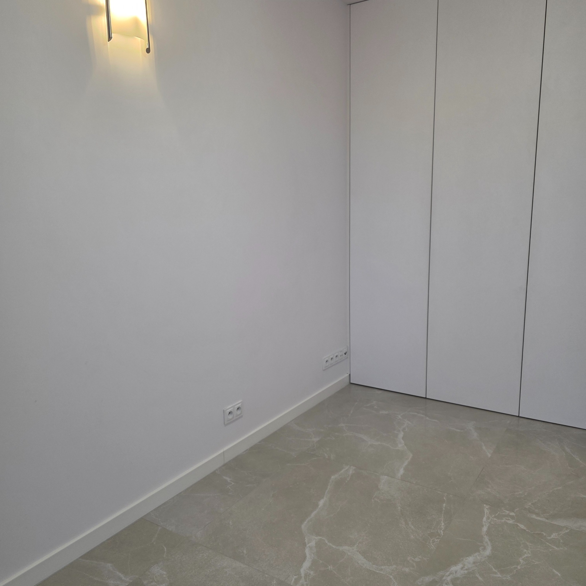 Minimalistyczne wnętrze: gładkie, białe ściany, marmurowe płytki podłogowe i wbudowana szafa z frontami bez uchwytów. Oświetlenie ścienne dodaje ciepła.