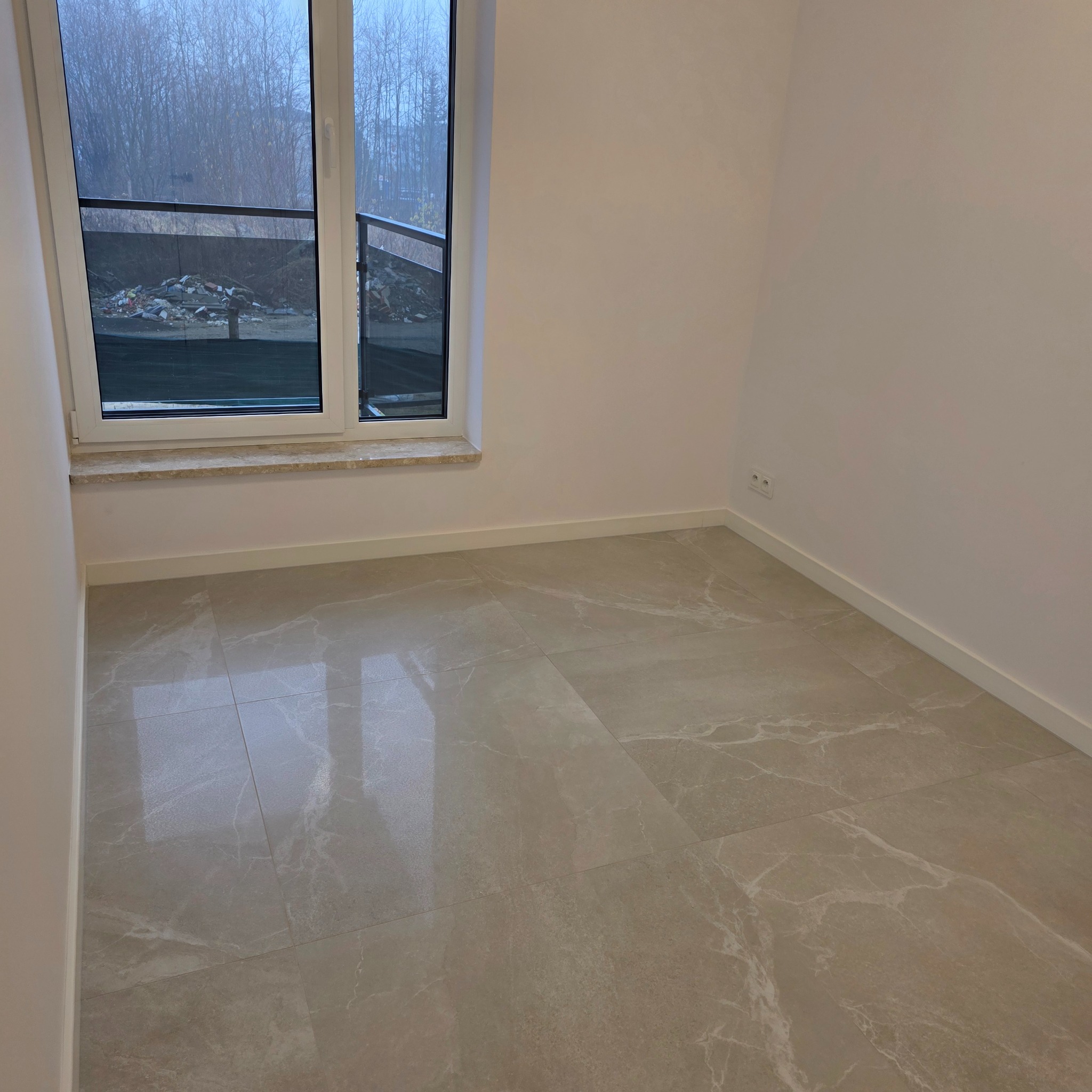 Jasne, marmurowe płytki podłogowe w pustym pokoju z oknem balkonowym. Widok na zewnątrz ukazuje drzewa i gruzowisko. Minimalistyczne wykończenie wnętrza.