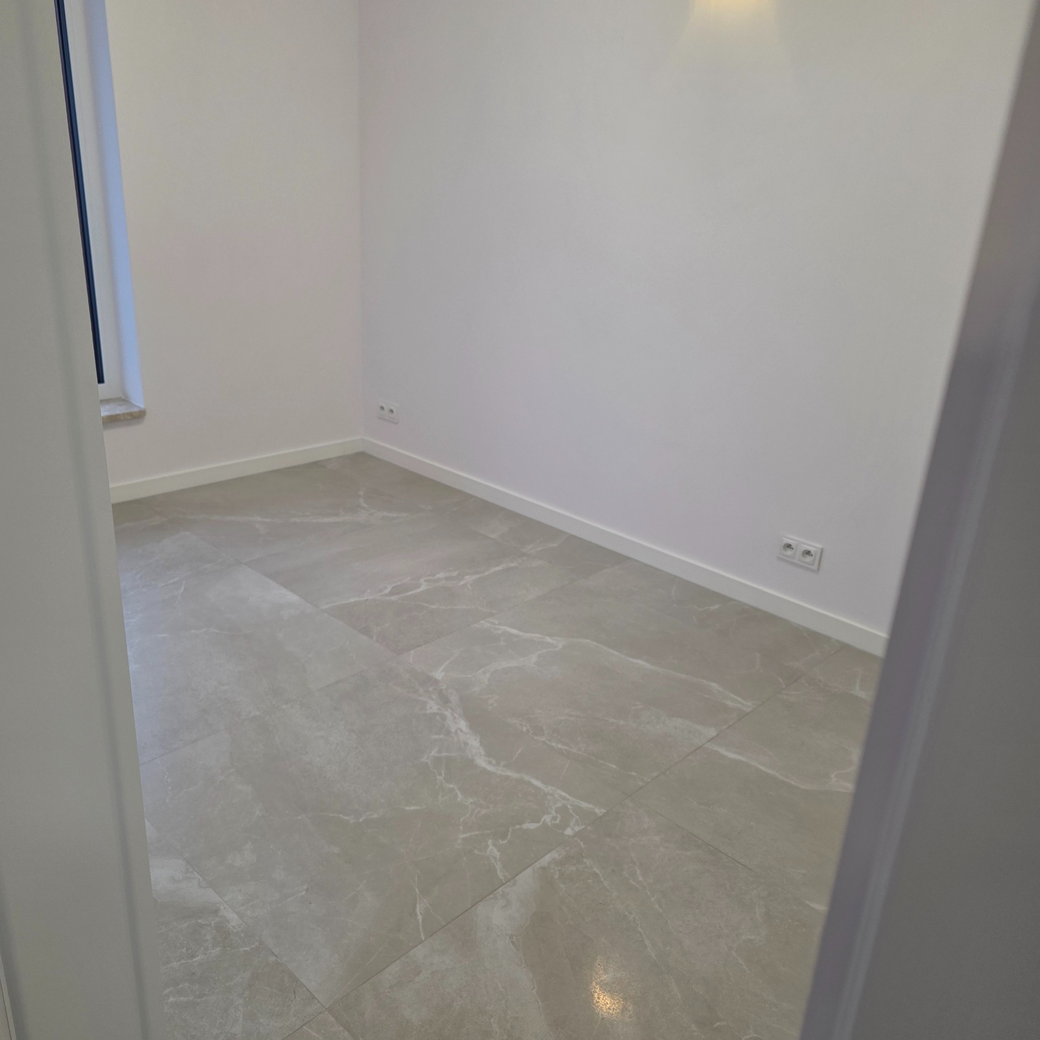 Wykończone wnętrze w Rydułtowach: szare płytki imitujące marmur na podłodze, białe ściany z gniazdkami. Widok zza futryny.