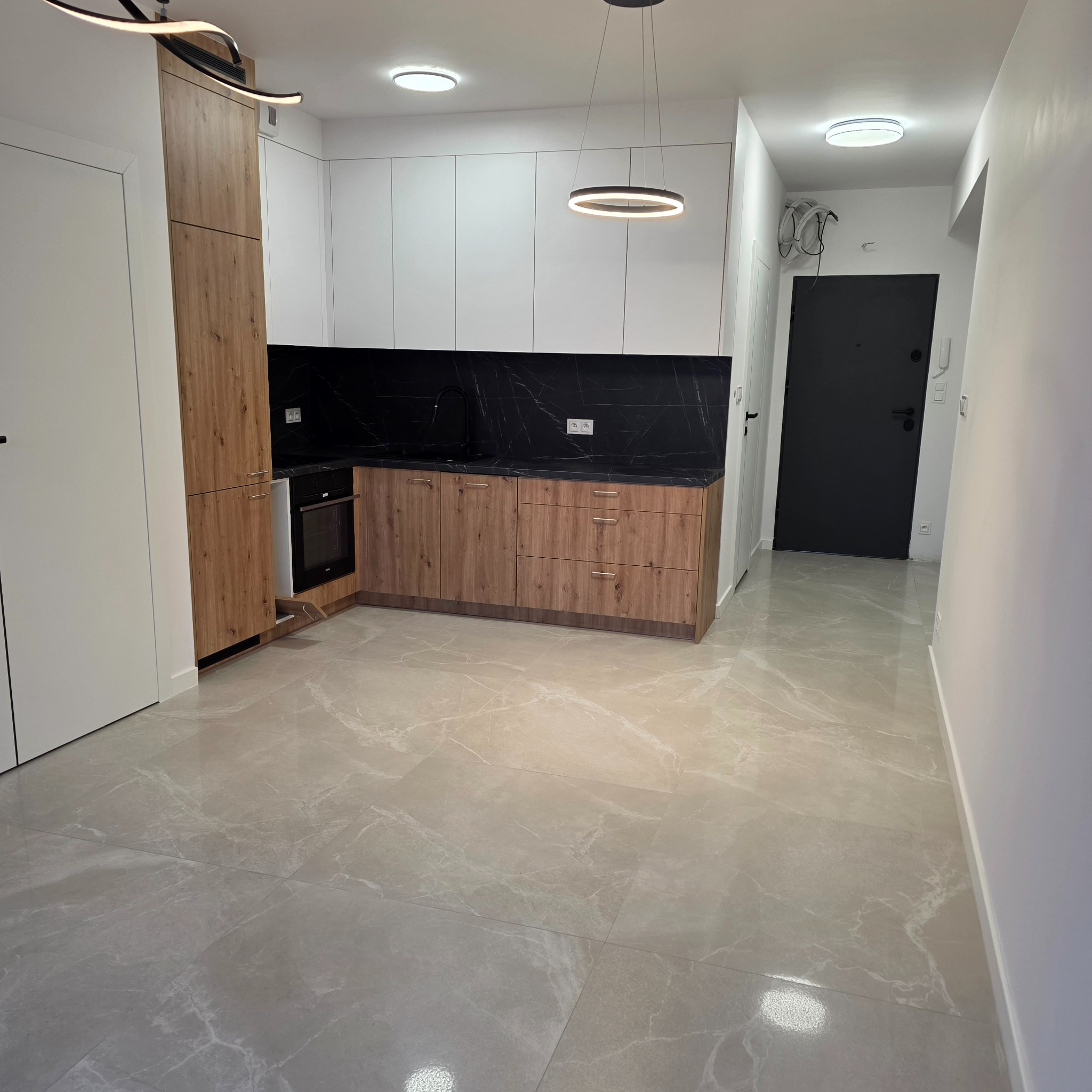 Nowoczesna kuchnia z drewnianymi szafkami i czarnym blatem, oświetlona okrągłą lampą. Jasne płytki podłogowe z marmurkowym wzorem. Minimalistyczny design.