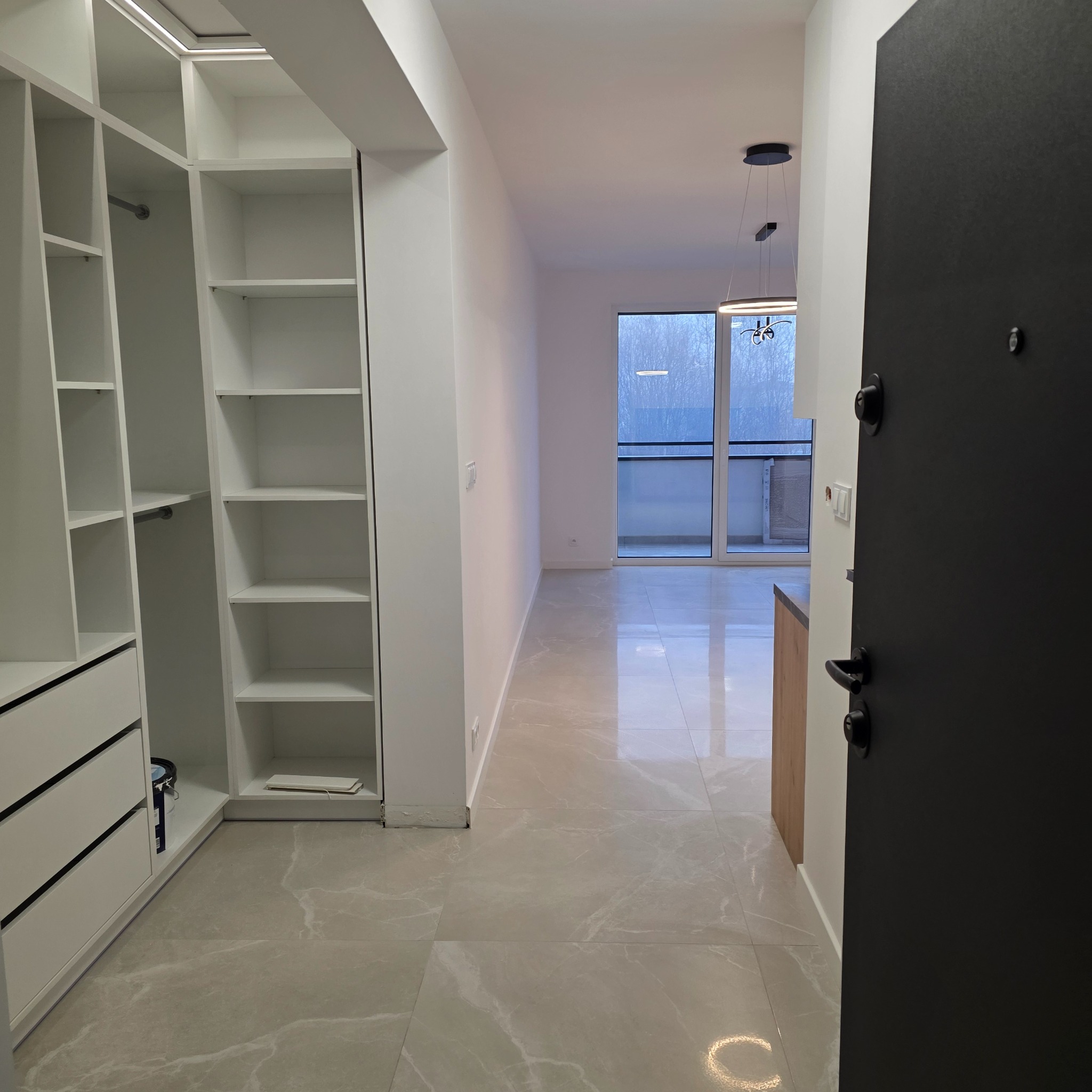 Nowoczesne wnętrze mieszkania z otwartą przestrzenią, widok z przedpokoju na salon z balkonem i zabudowaną szafą wnękową z białymi półkami.