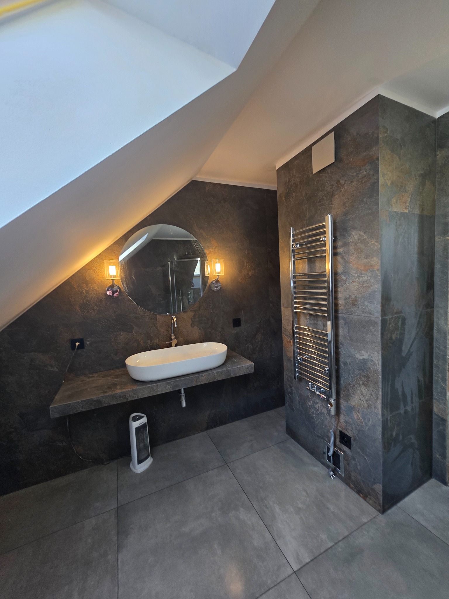 Nowoczesna łazienka na poddaszu z owalną umywalką, okrągłym lustrem i grzejnikiem drabinkowym na tle ciemnych płytek imitujących kamień.