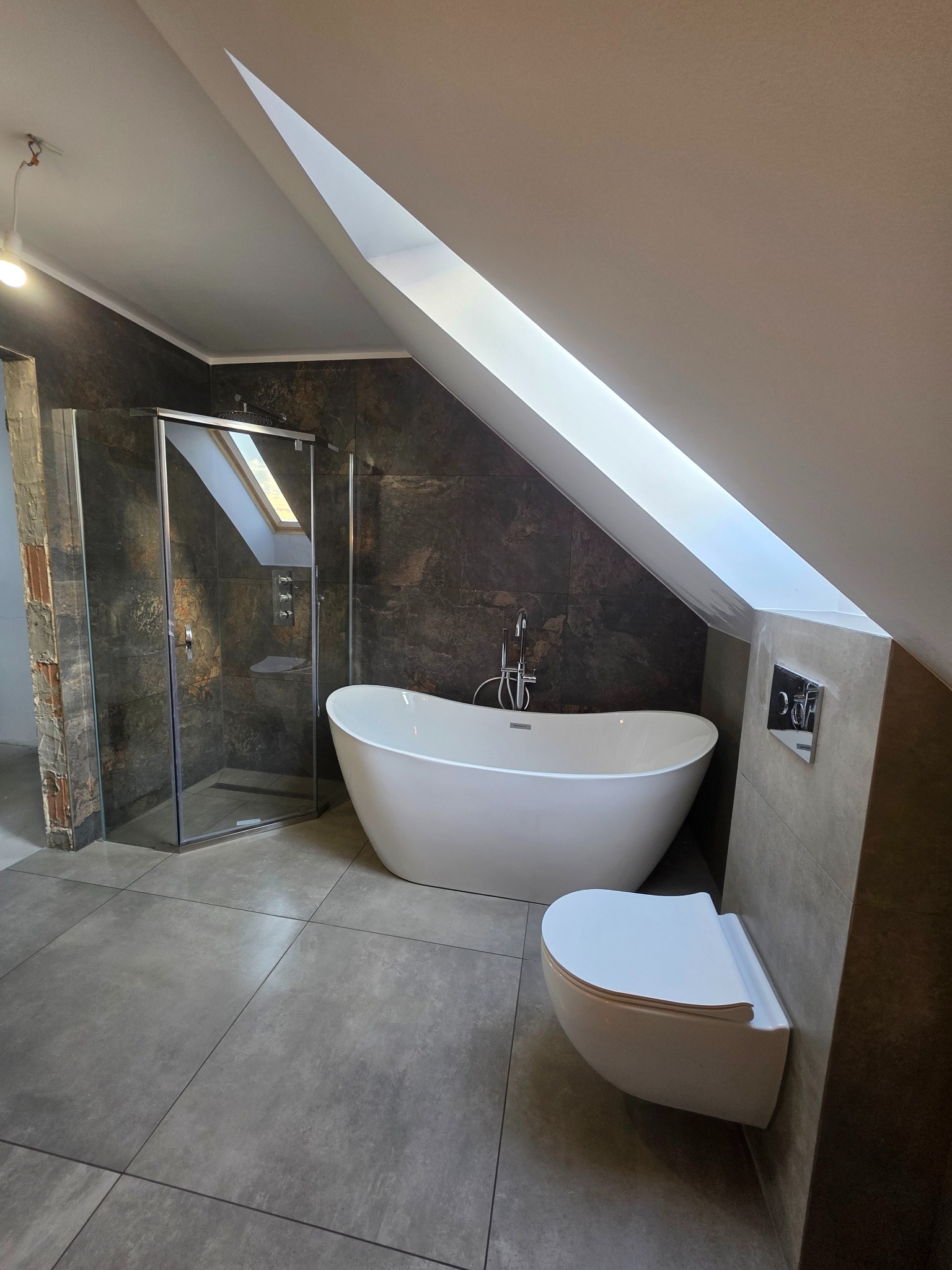 Nowoczesna łazienka na poddaszu z wolnostojącą wanną, kabiną prysznicową i podwieszaną toaletą. Ciemne płytki imitujące kamień kontrastują z bielą ceramiki.