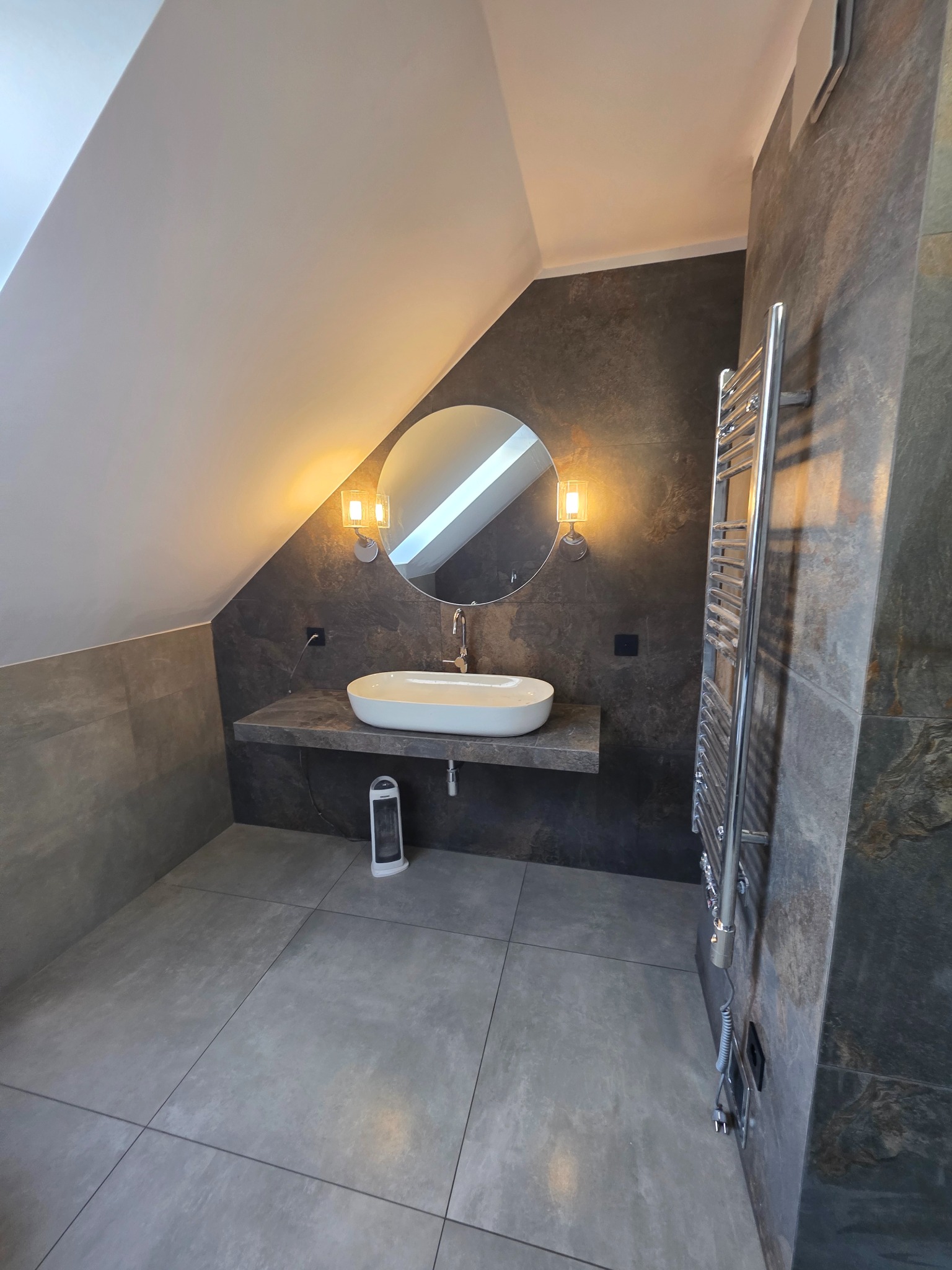 Nowoczesna łazienka na poddaszu w Rydułtowach. Umywalka nablatowa, okrągłe lustro z oświetleniem, grzejnik drabinkowy i szare płytki tworzą spójną całość.