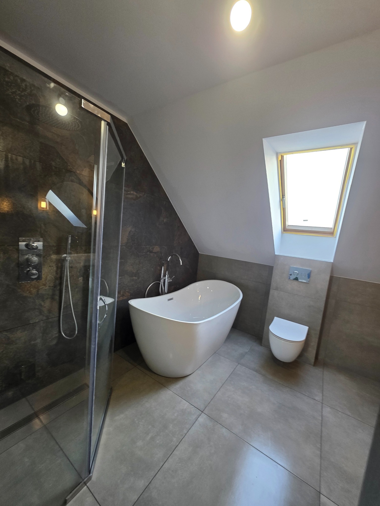 Nowoczesna łazienka na poddaszu z kabiną prysznicową, wolnostojącą wanną i toaletą. Ciemne płytki kontrastują z bielą armatury i jasnym oknem dachowym.