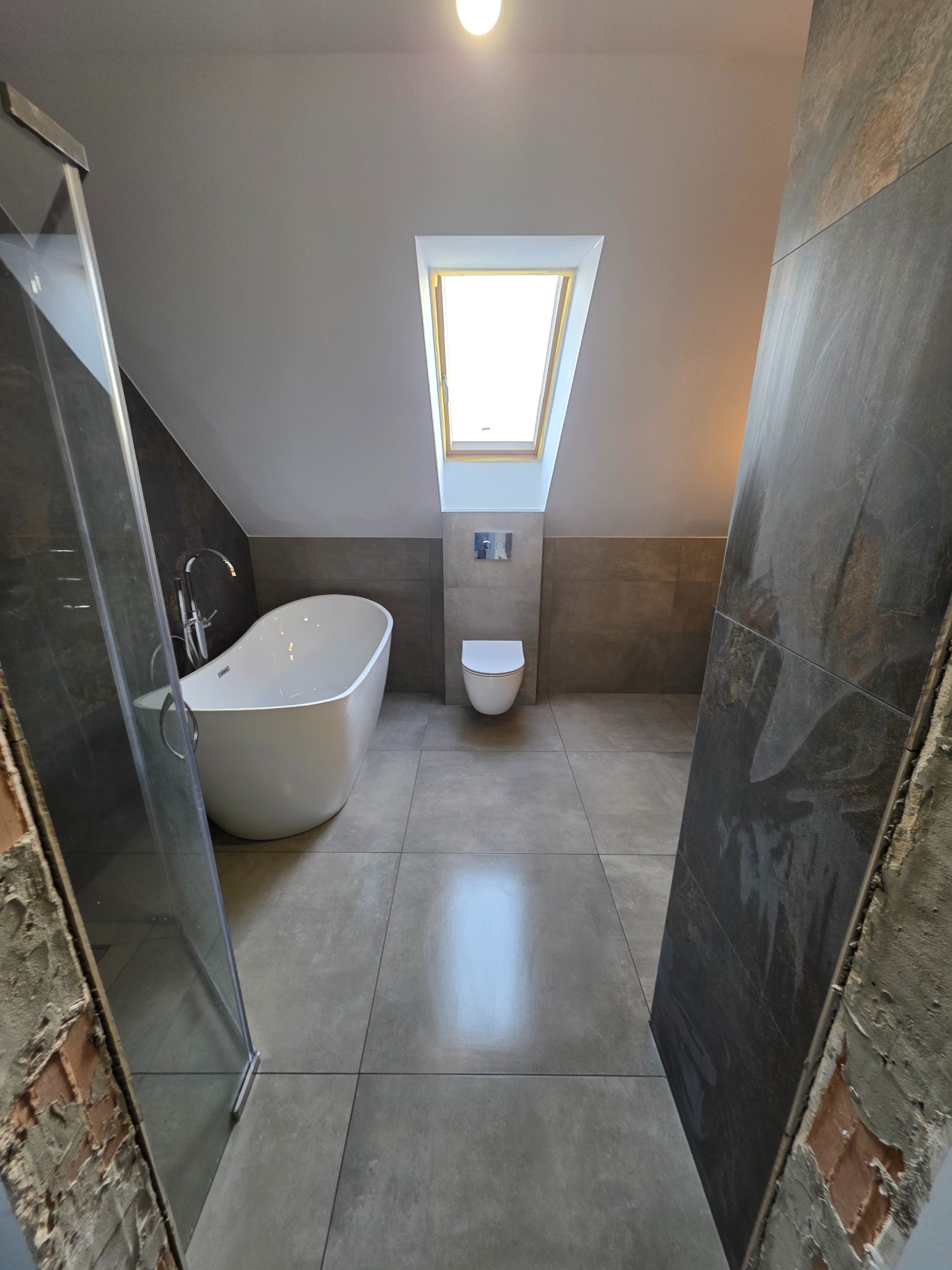 Nowoczesna łazienka na poddaszu z oknem dachowym, wolnostojącą wanną i kabiną prysznicową. Szare płytki na podłodze i ścianach, minimalistyczna toaleta.