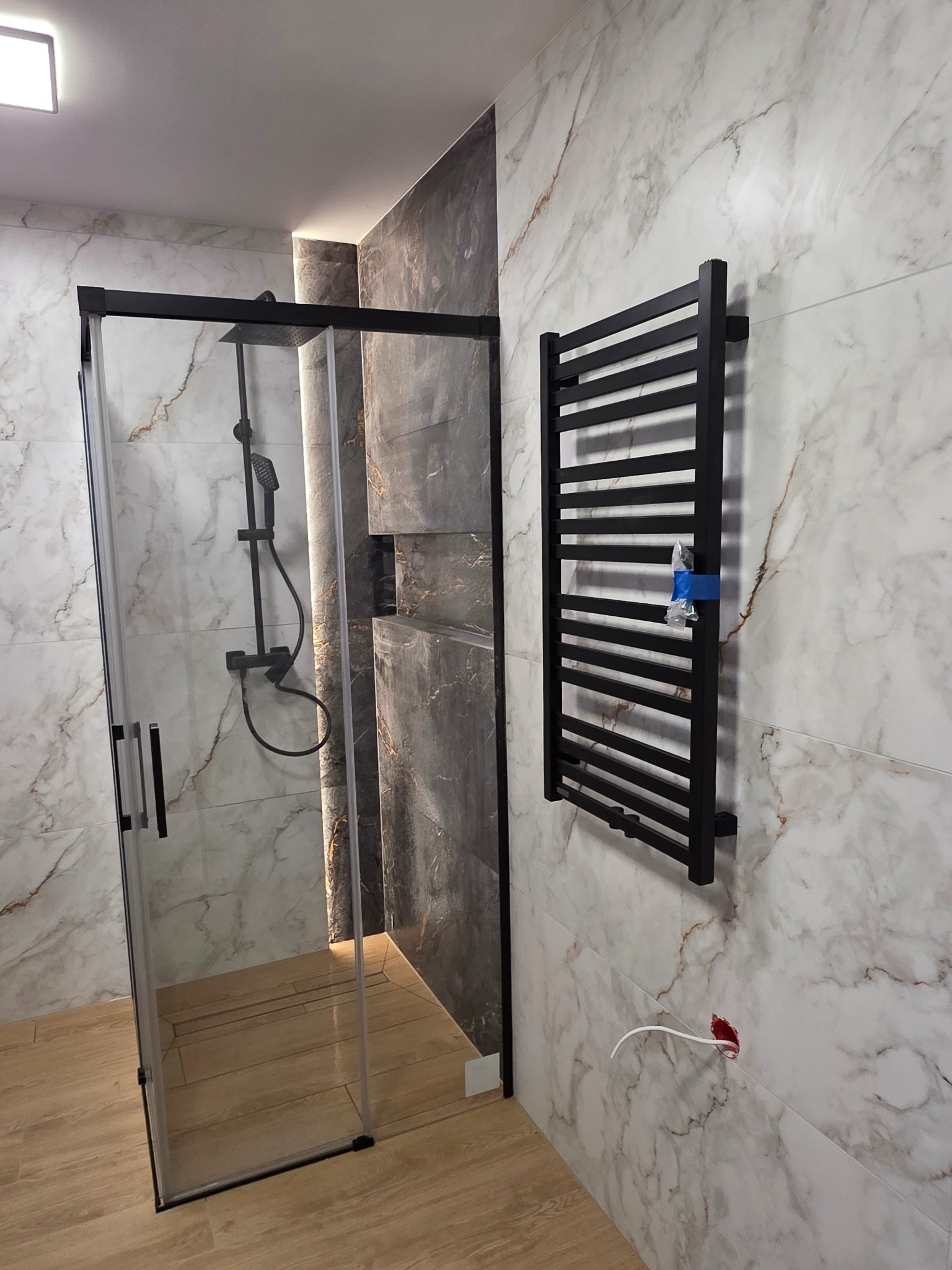 Wykończona łazienka z kabiną prysznicową typu walk-in z czarnymi profilami, grzejnikiem drabinkowym i marmurowymi płytkami. Widoczne niedokończone podłączenie elektryczne.