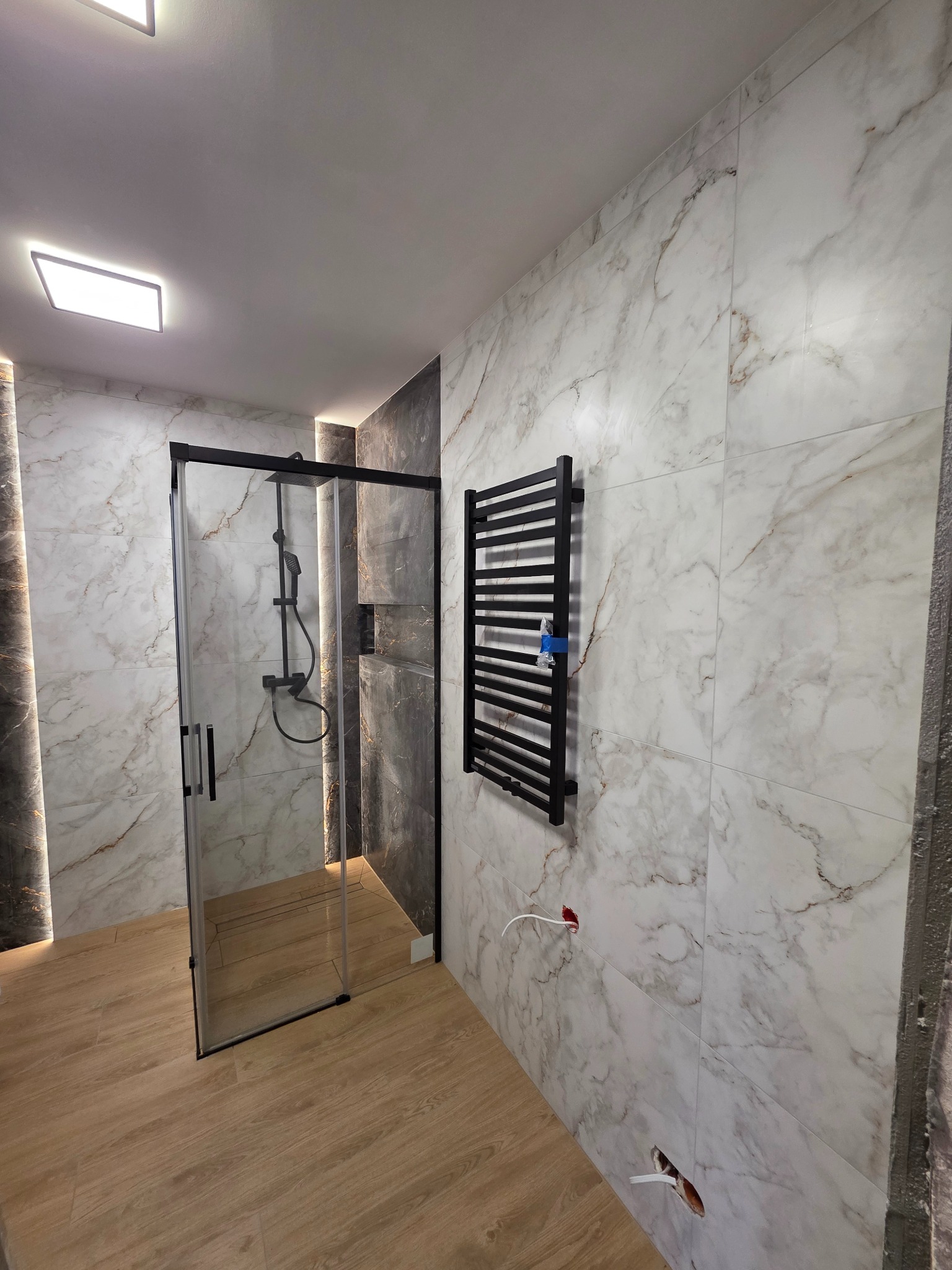 Nowoczesna łazienka po remoncie: kabina prysznicowa z czarnymi profilami, grzejnik drabinkowy, marmurowe płytki ścienne i podłoga imitująca drewno. Widoczne punkty elektryczne.