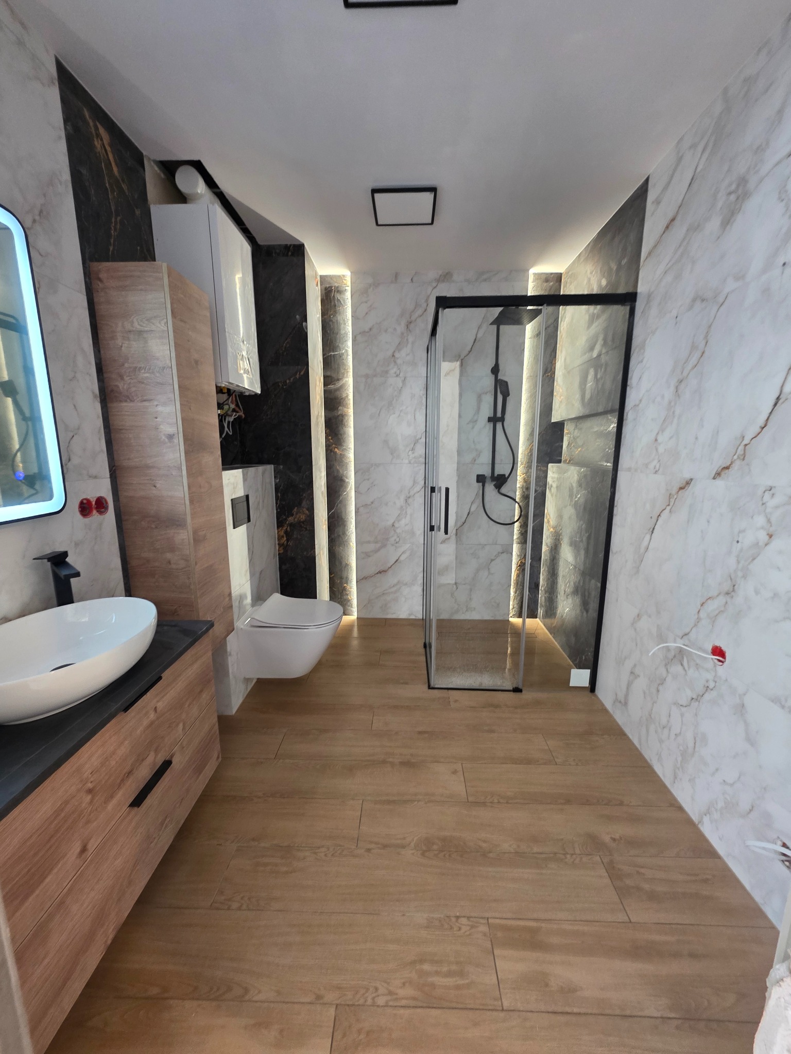 Nowoczesna łazienka po remoncie: drewnopodobne płytki, kabina prysznicowa z czarnymi profilami, umywalka nablatowa i marmurowe płytki z podświetleniem LED.