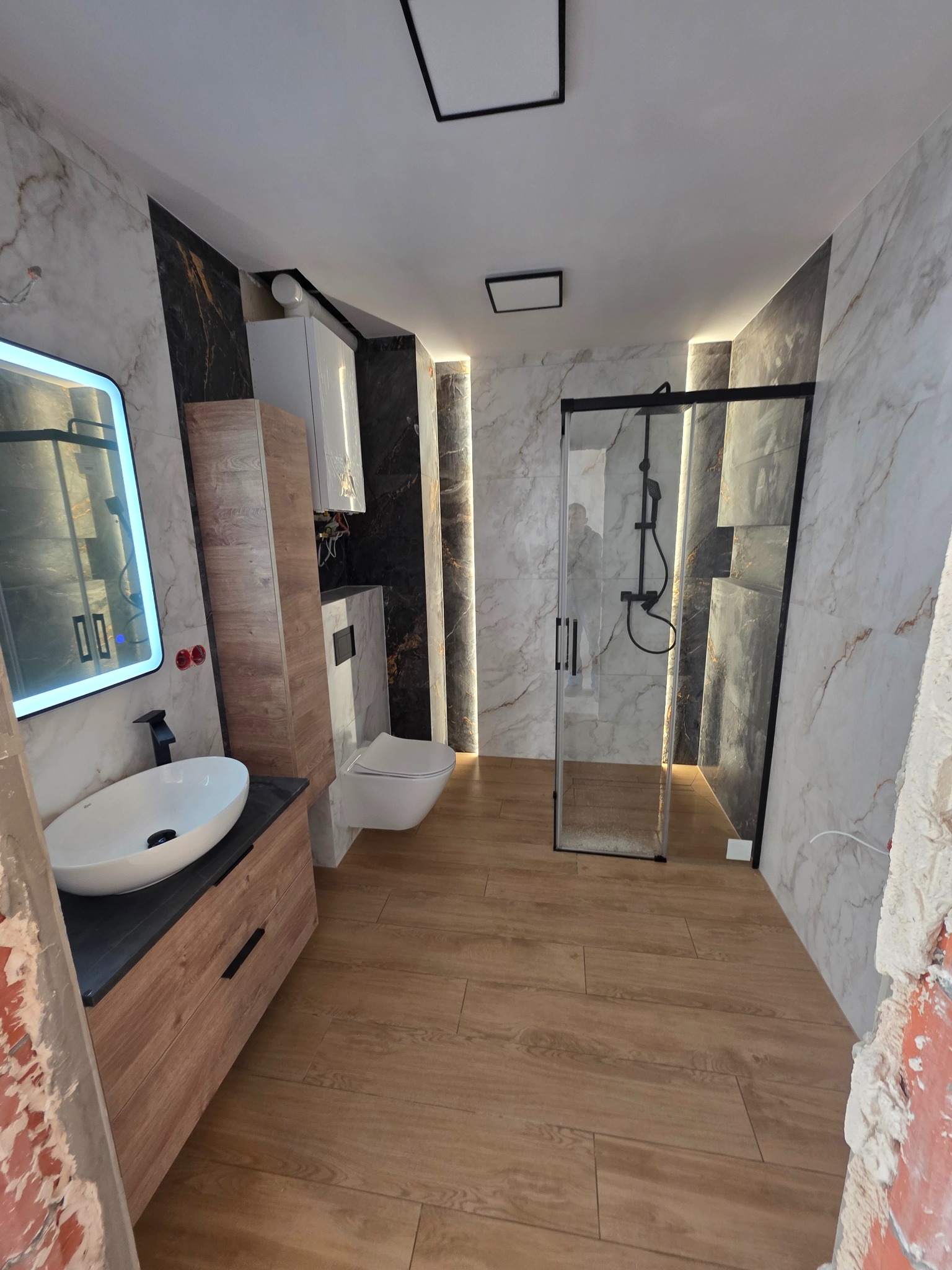 Nowoczesna łazienka z drewnianą podłogą, marmurowymi płytkami i podświetlaną kabiną prysznicową. Umywalka z podświetlanym lustrem i drewniana szafka.