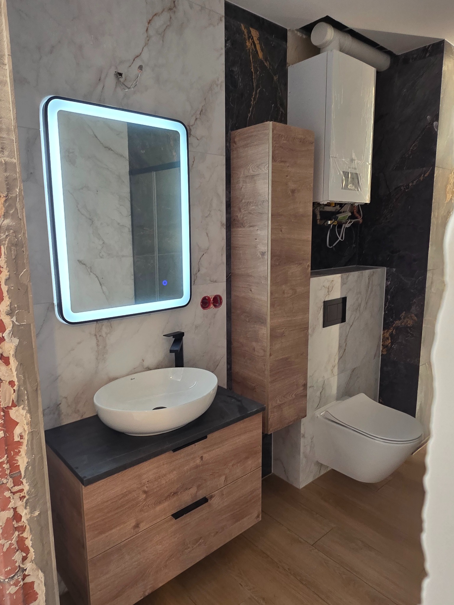 Nowoczesna łazienka z podświetlanym lustrem, umywalką na szafce z drewnianym dekorem, wiszącą toaletą i marmurowymi płytkami na ścianach. Widoczny piec gazowy.