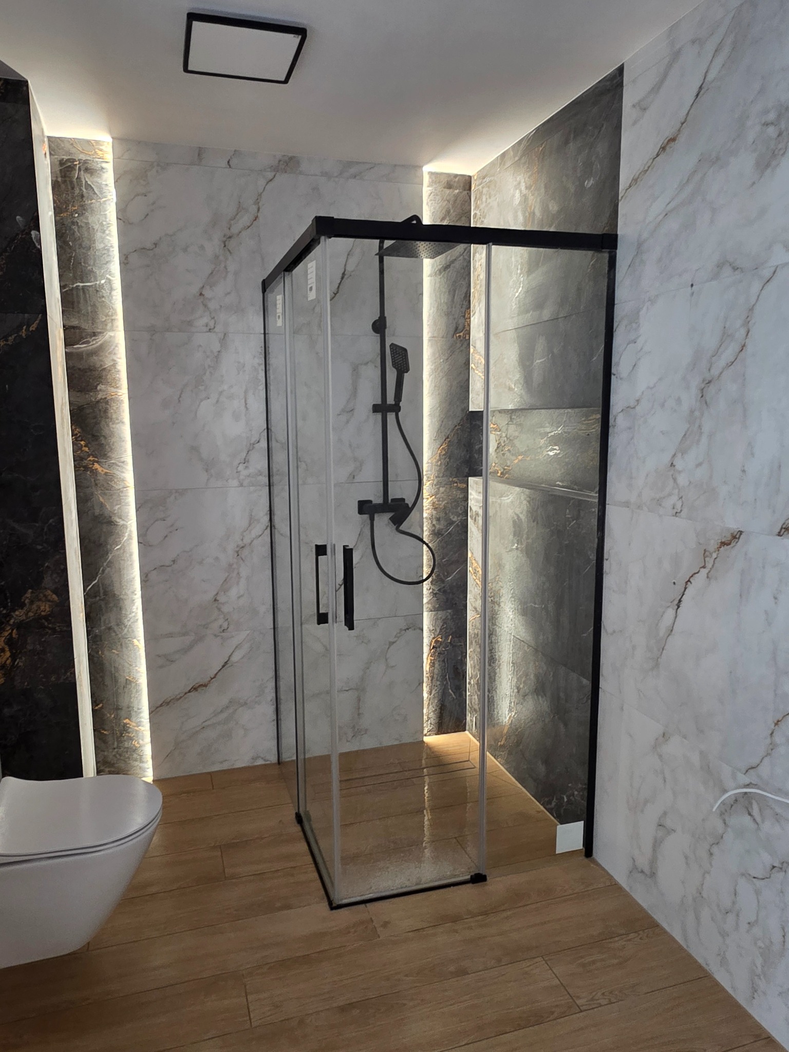 Nowoczesna łazienka z kabiną prysznicową typu walk-in, marmurowe płytki na ścianach z podświetleniem LED, drewnopodobna podłoga i wisząca toaleta.