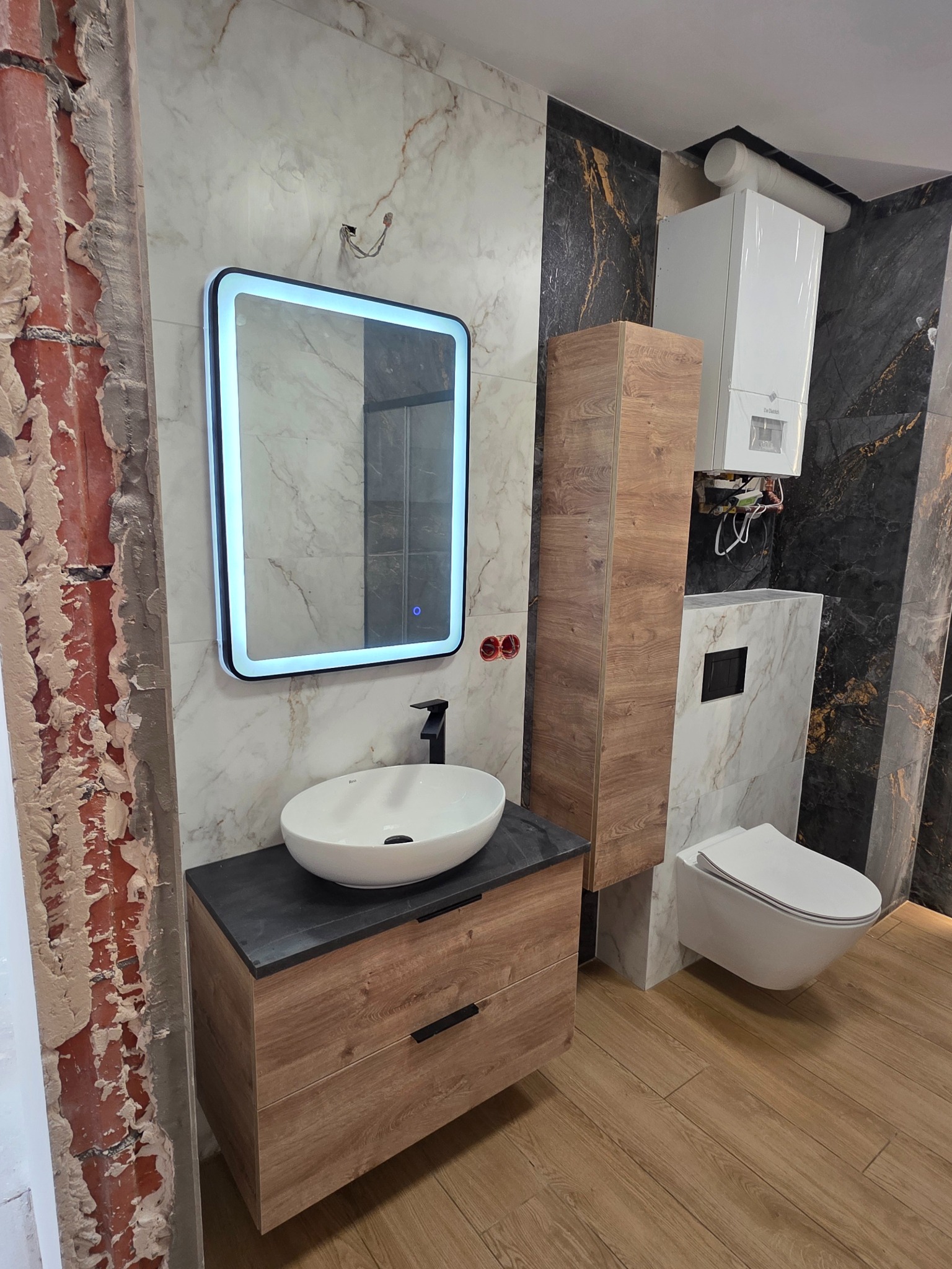 Nowoczesna łazienka z umywalką nablatową, podświetlanym lustrem i płytkami imitującymi marmur. Fragment ściany w trakcie wykończenia.