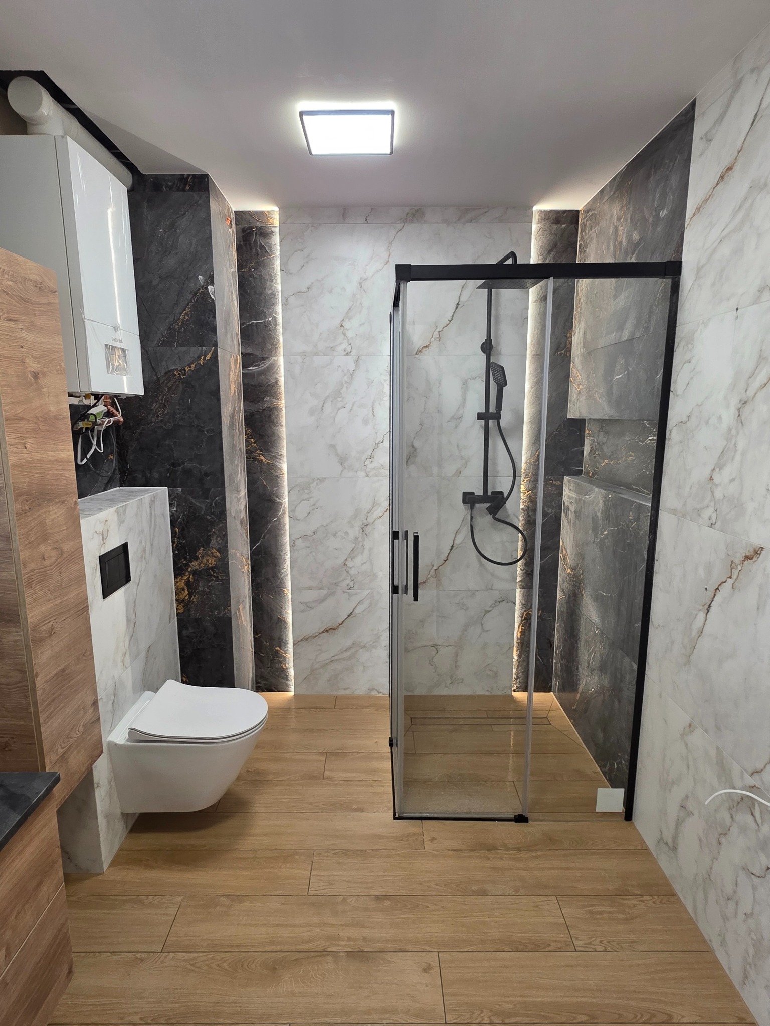 Wykończona łazienka z kabiną prysznicową typu walk-in, podwieszaną toaletą i płytkami imitującymi marmur w odcieniach bieli i szarości. Oświetlenie LED podkreśla pionowe linie.