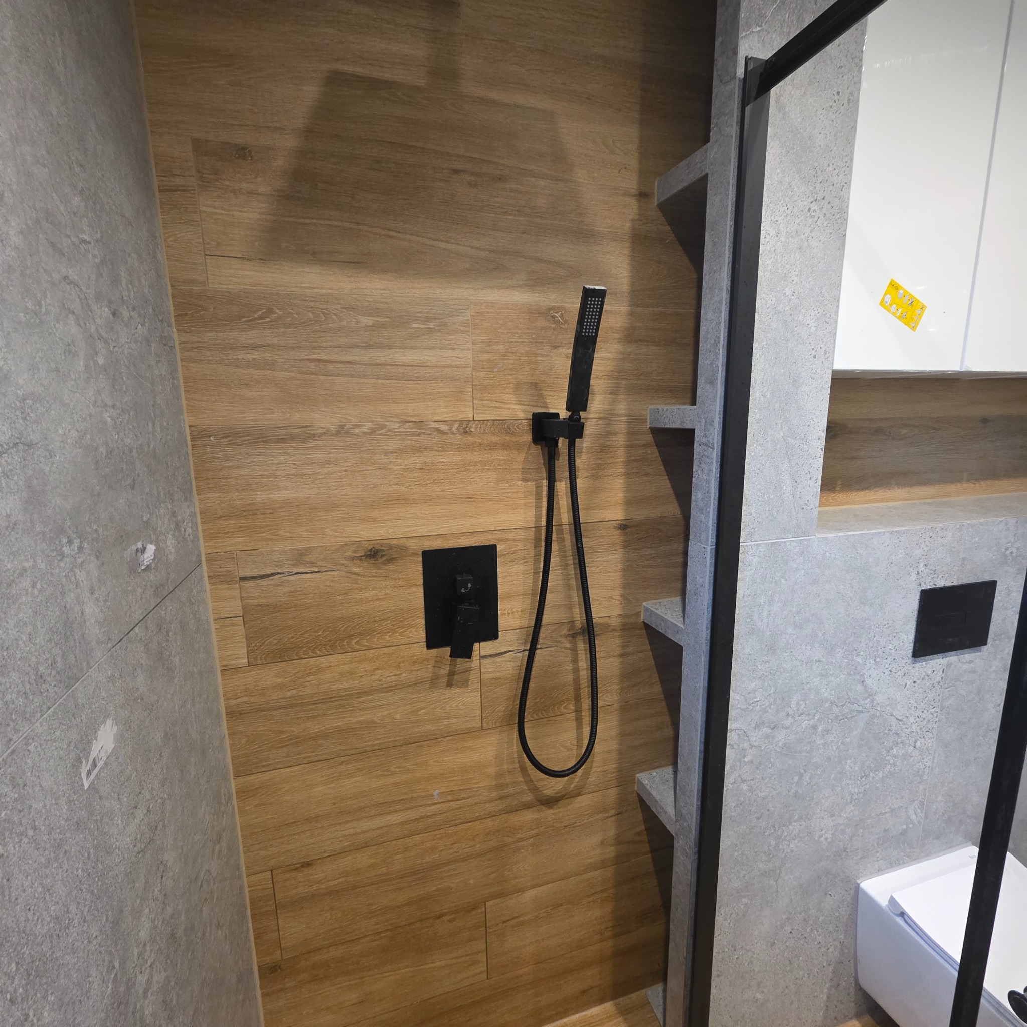 Nowoczesna kabina prysznicowa z drewnopodobnymi płytkami i czarną armaturą. Minimalistyczny design z szarymi półkami i elementami wykończenia.