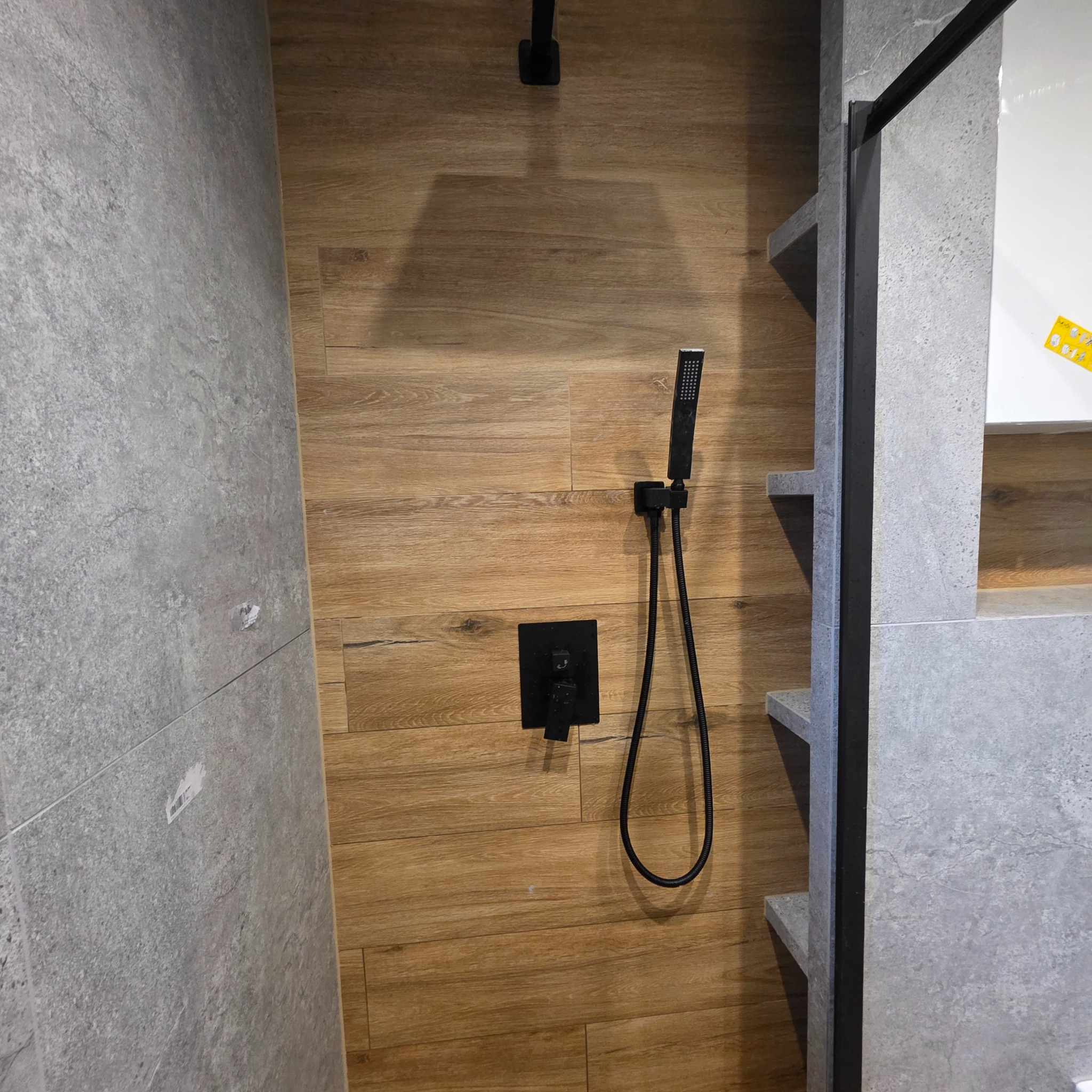 Nowoczesna kabina prysznicowa z drewnopodobnymi i szarymi płytkami, czarna armatura, półki wnękowe. Minimalistyczny design łazienki.