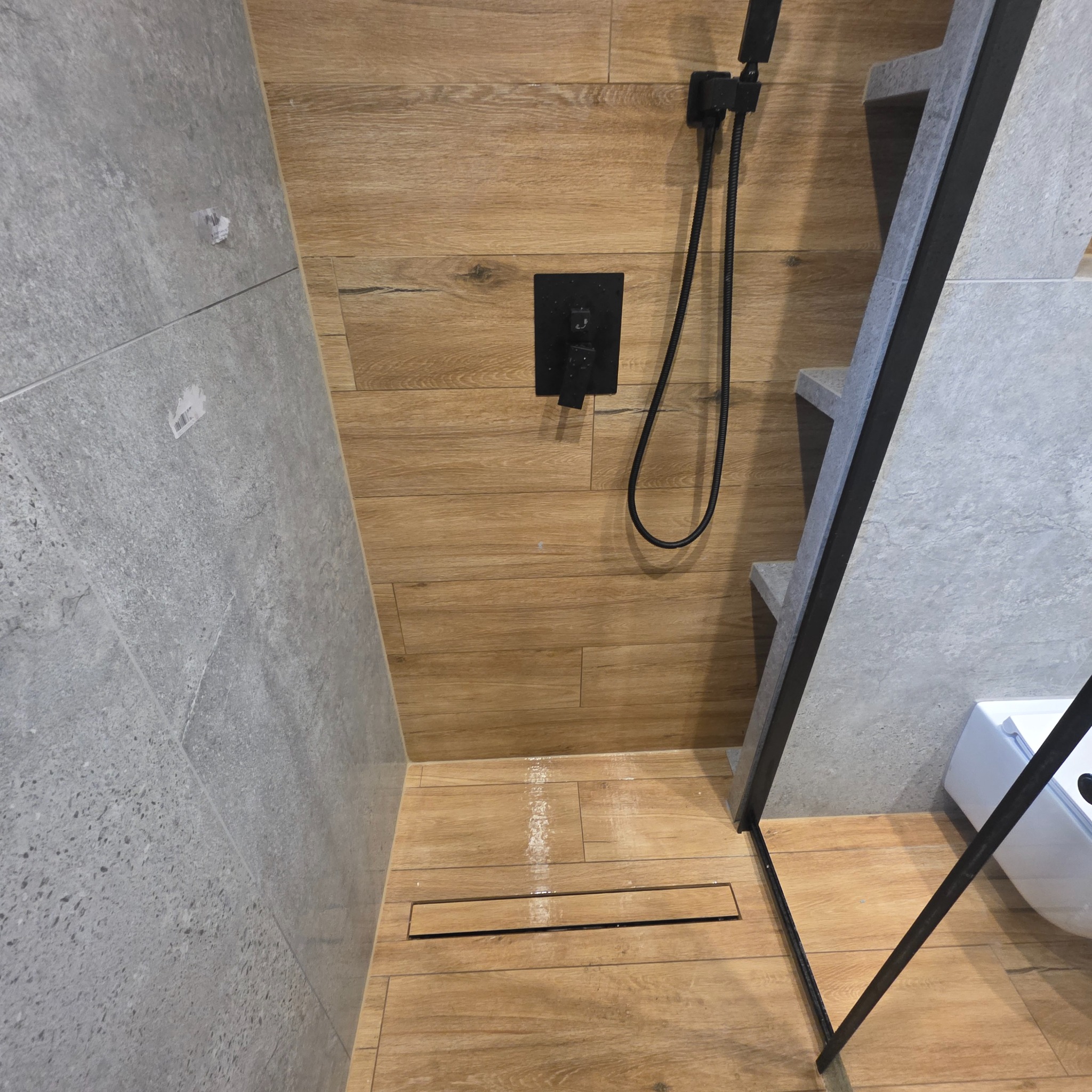 Nowoczesna kabina prysznicowa z drewnopodobnymi płytkami i czarną armaturą. Uwagę zwraca liniowy odpływ i nietypowe schodki z szarej płytki.