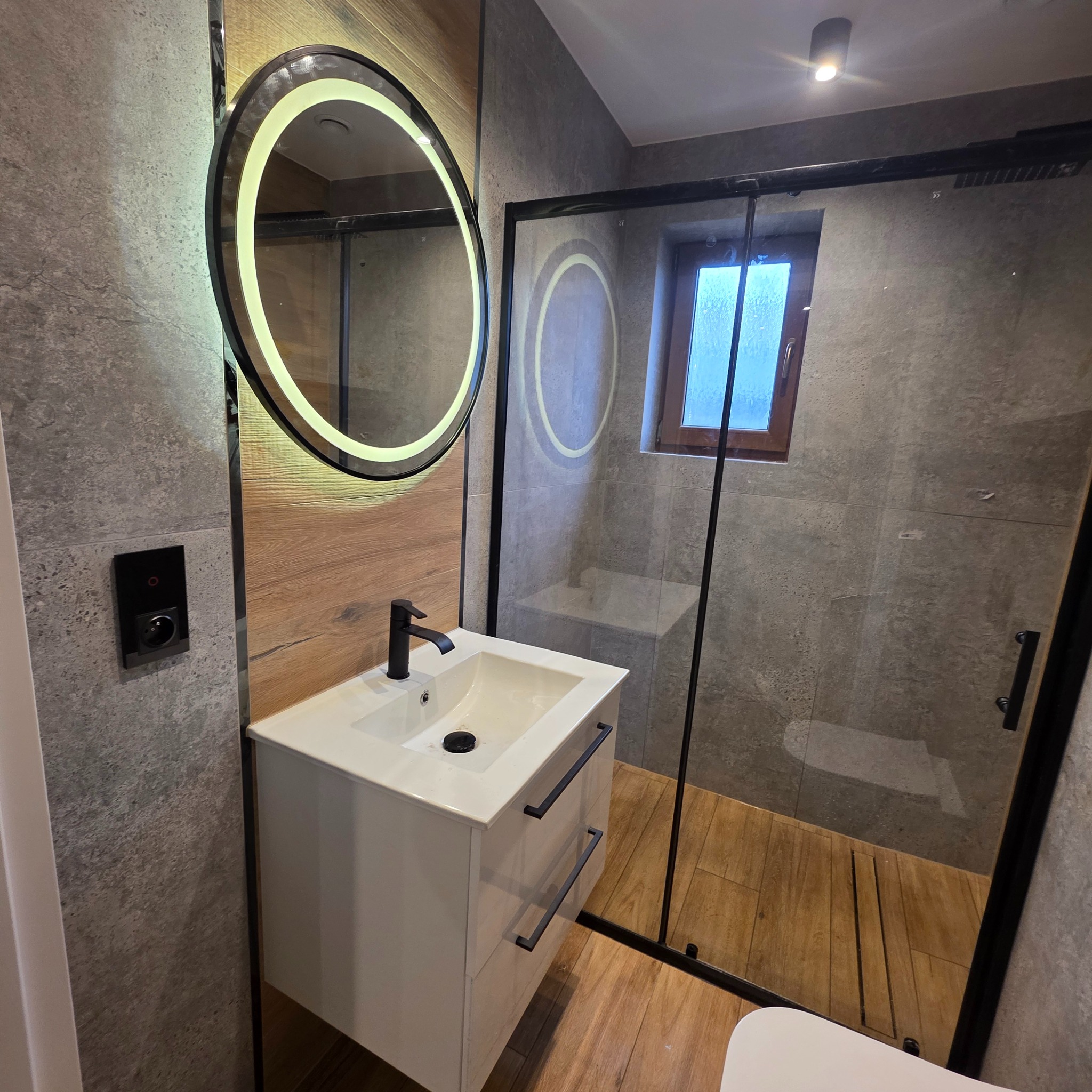 Nowoczesna łazienka z drewnianymi akcentami: okrągłe lustro z podświetleniem LED, biała szafka z umywalką i czarną armaturą, kabina prysznicowa z czarną ramą.