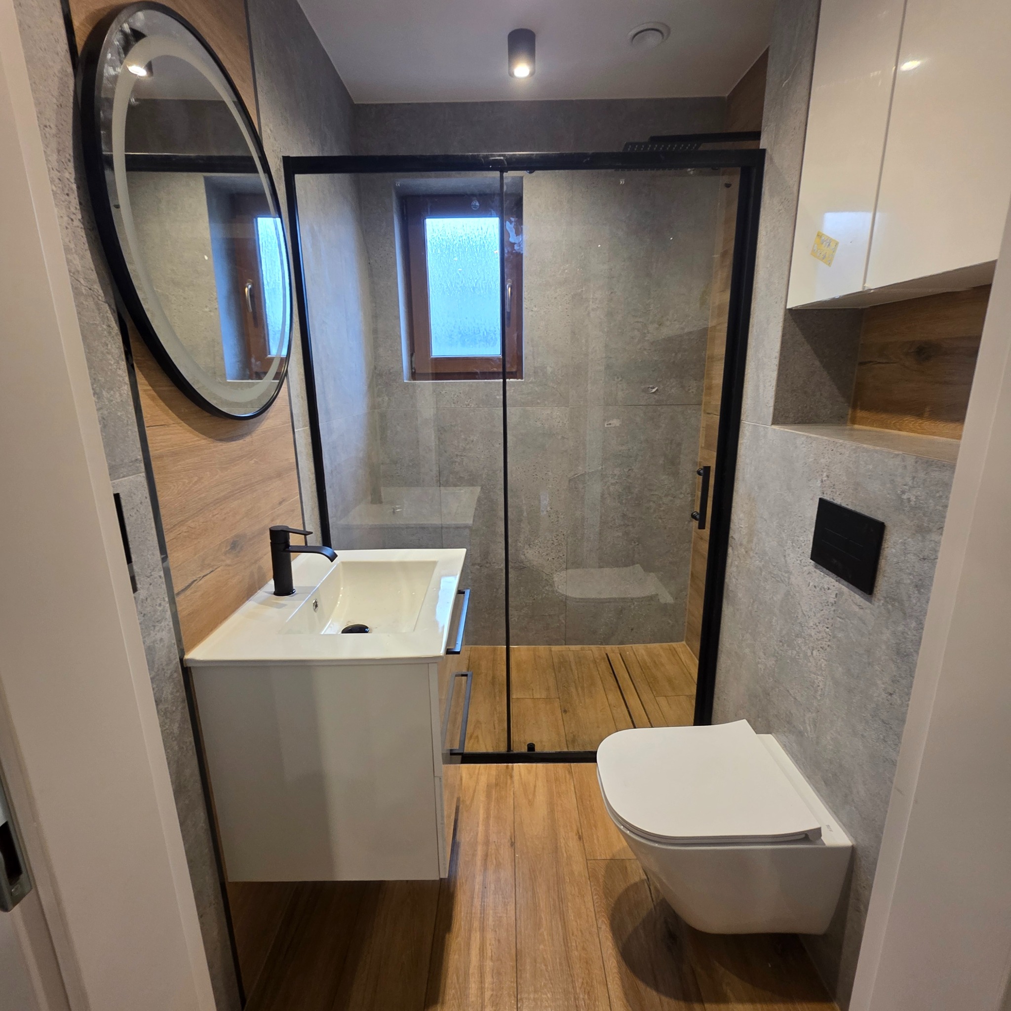 Nowoczesna łazienka z kabiną prysznicową typu walk-in, umywalką na szafce i podwieszaną toaletą. Uwagę zwraca okrągłe lustro z podświetleniem i drewnopodobne płytki.