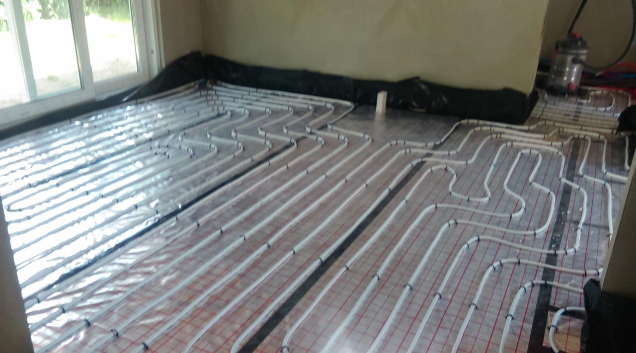 Białe rury ogrzewania podłogowego ułożone na folii izolacyjnej z czerwoną siatką w pomieszczeniu z oknem i ścianami w kolorze beżowym. Widoczny fragment instalacji.
