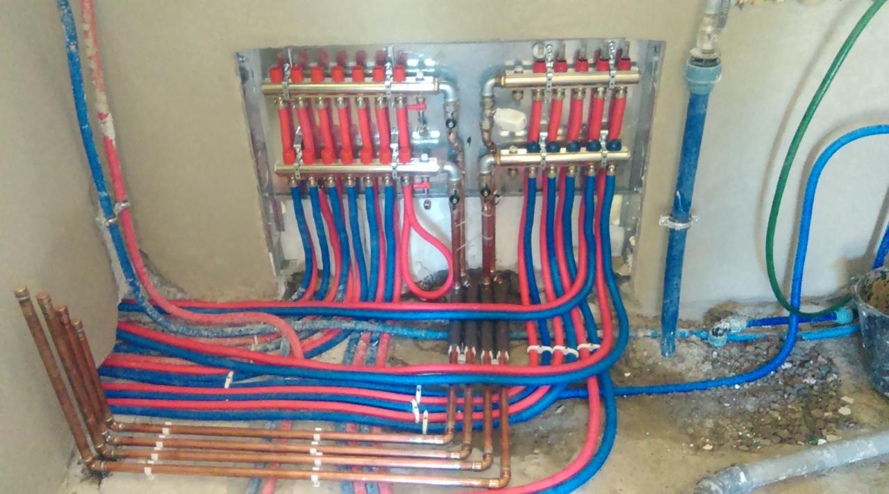 Rozdzielacz instalacji grzewczej podłączony rurami w kolorach niebieskim i czerwonym. Widoczne miedziane rury i fragment ściany. Instalacja w trakcie montażu.